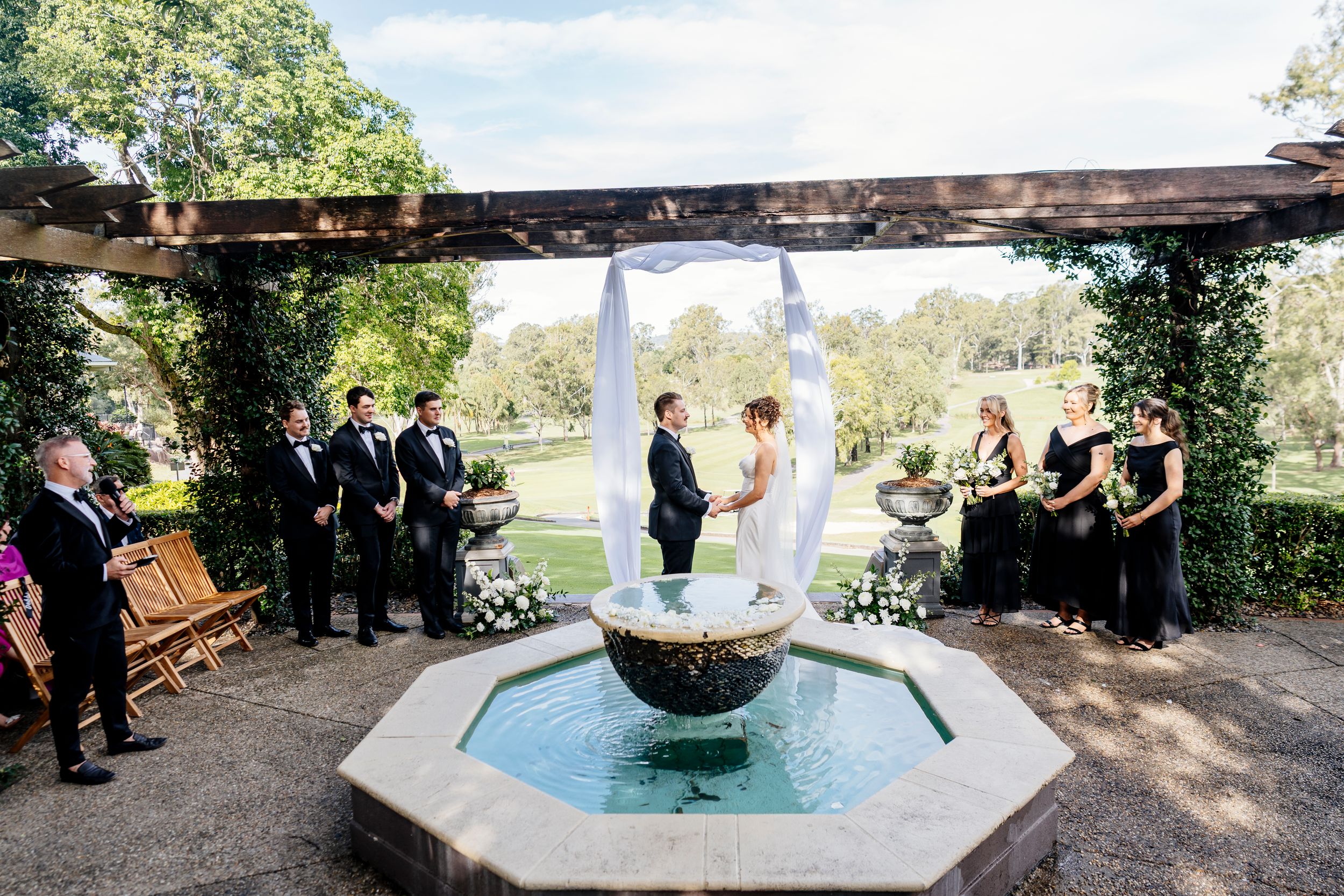 Outdoor wedding ceremony by a stone fountain under a pergola, with bride & groom exchanging vows surrounded by wedding party Hillstone