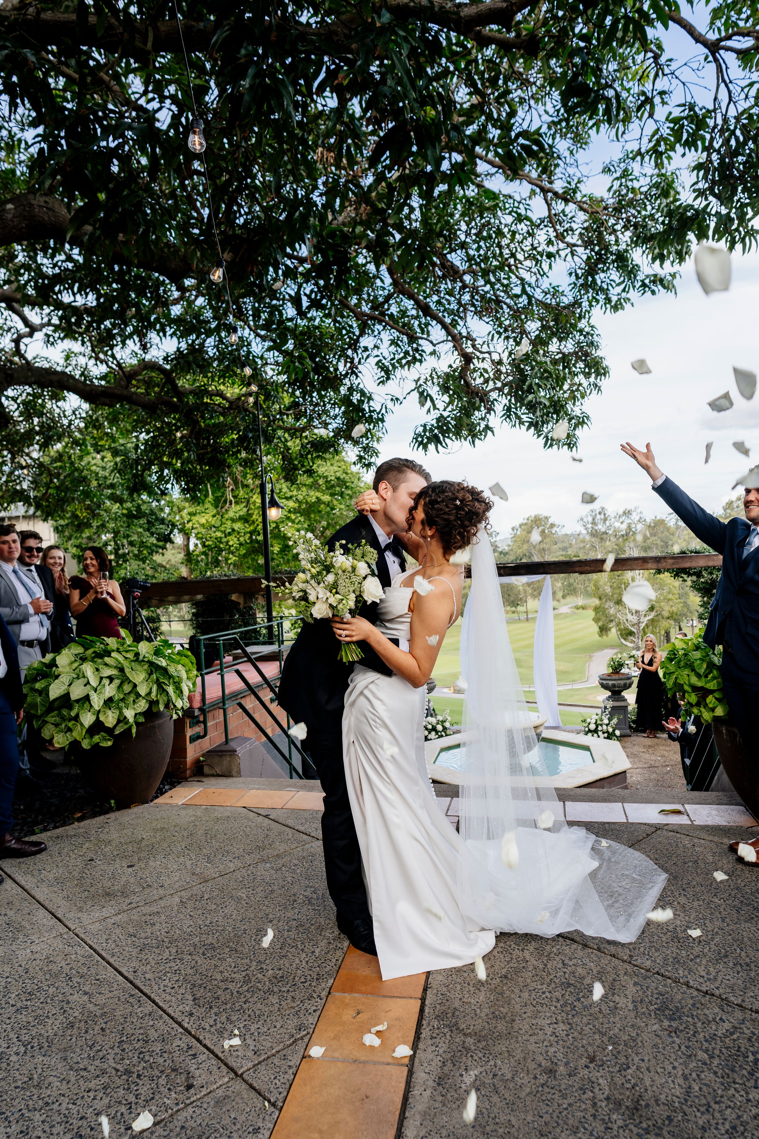 Bride and groom share first kiss outdoors under tree with white rose petals tossed by guests at elegant wedding ceremony Hillstone St Lucia