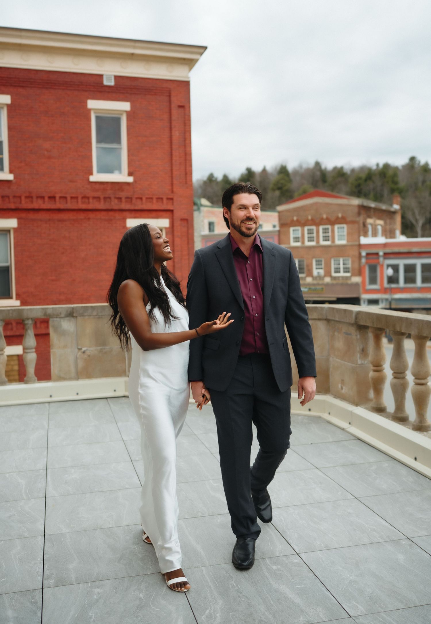 A smiling interracial couple walks together on a rooftop terrace, woman in white jumpsuit and man in dark suit.