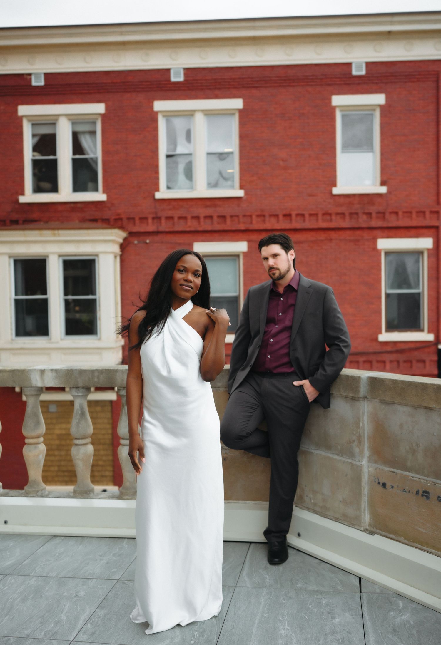 Interracial couple posing on rooftop, woman in white halter gown, man in gray suit, red brick building backdrop.