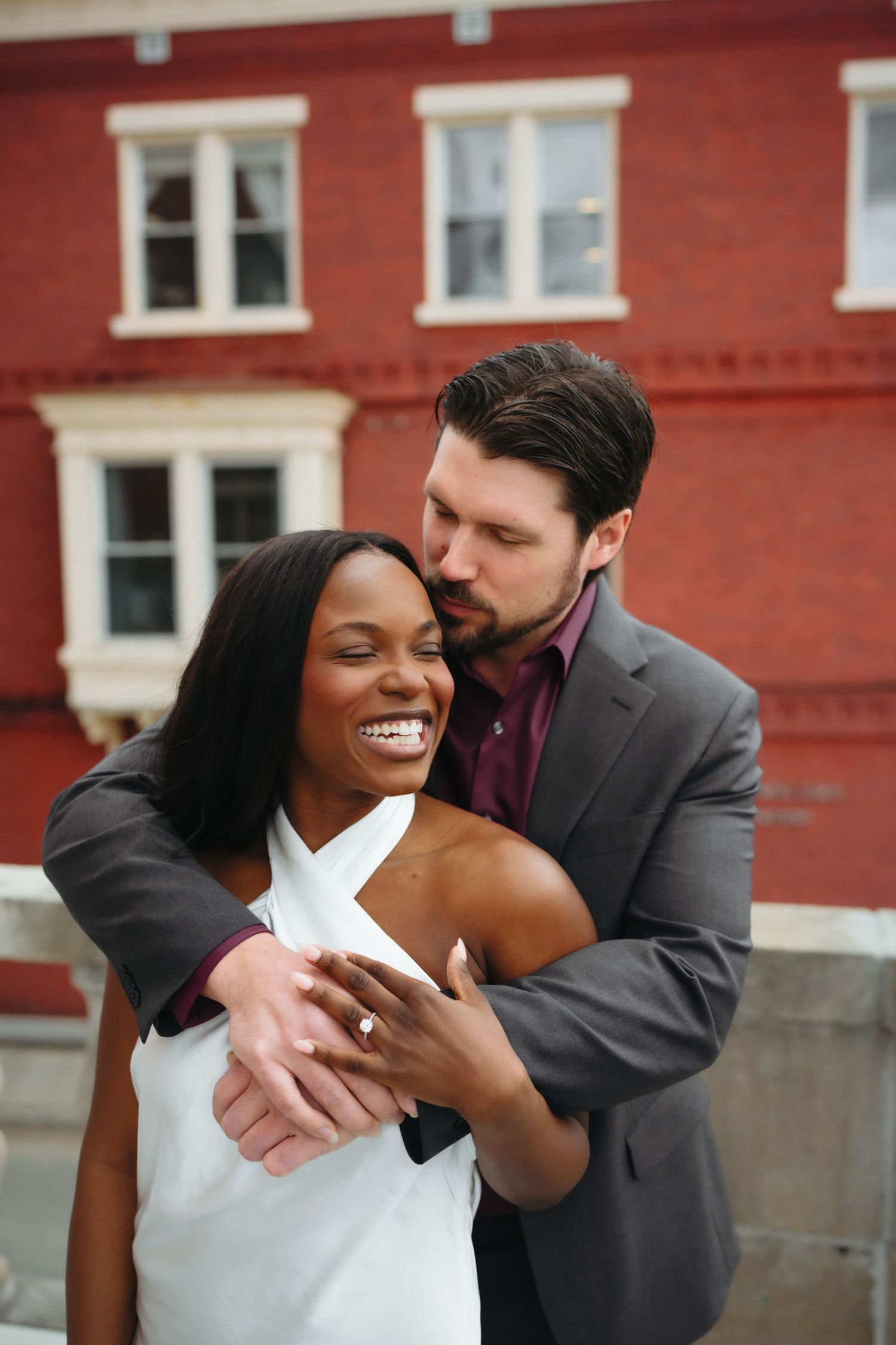 Interracial couple embracing outdoors, woman in white dress smiling, man in gray suit kissing her cheek near red brick building.