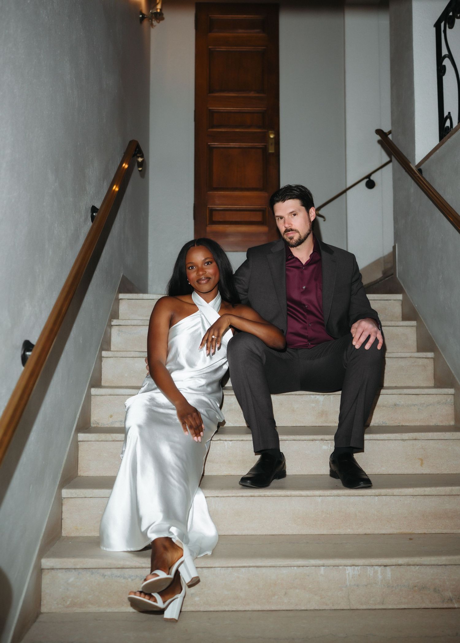 Elegant interracial couple sitting on marble staircase, woman in white satin dress, man in charcoal suit.