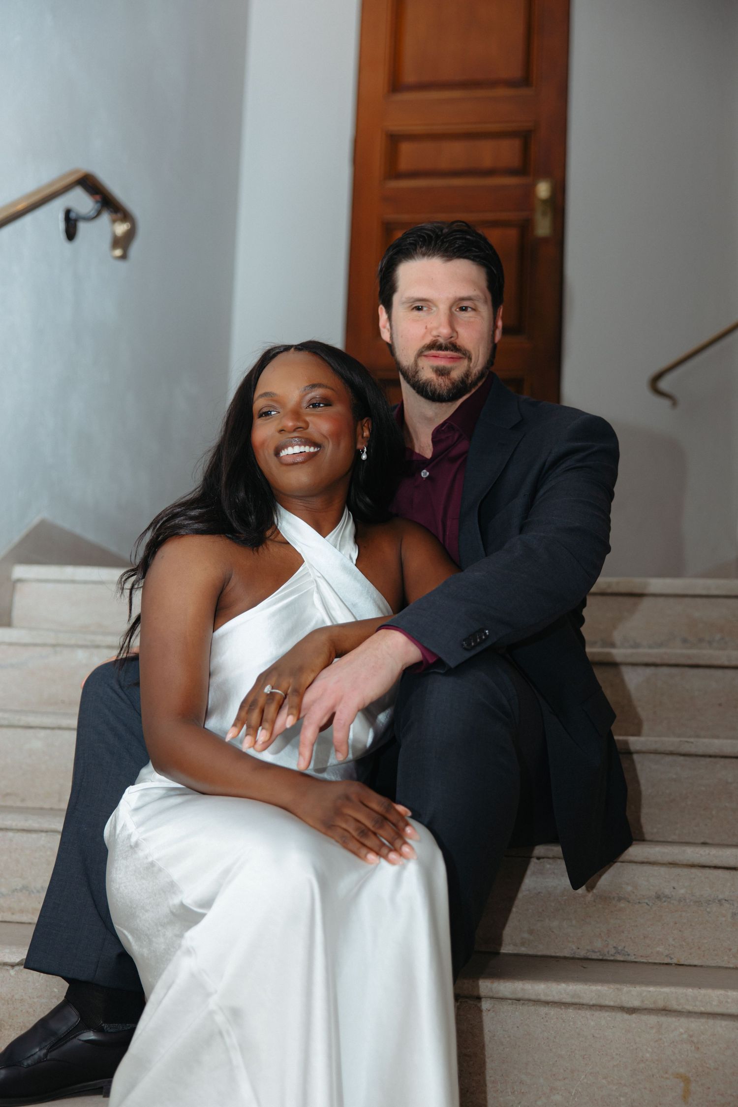 A smiling interracial couple in formal attire sits together on marble stairs, the woman in a white dress and man in a dark suit.