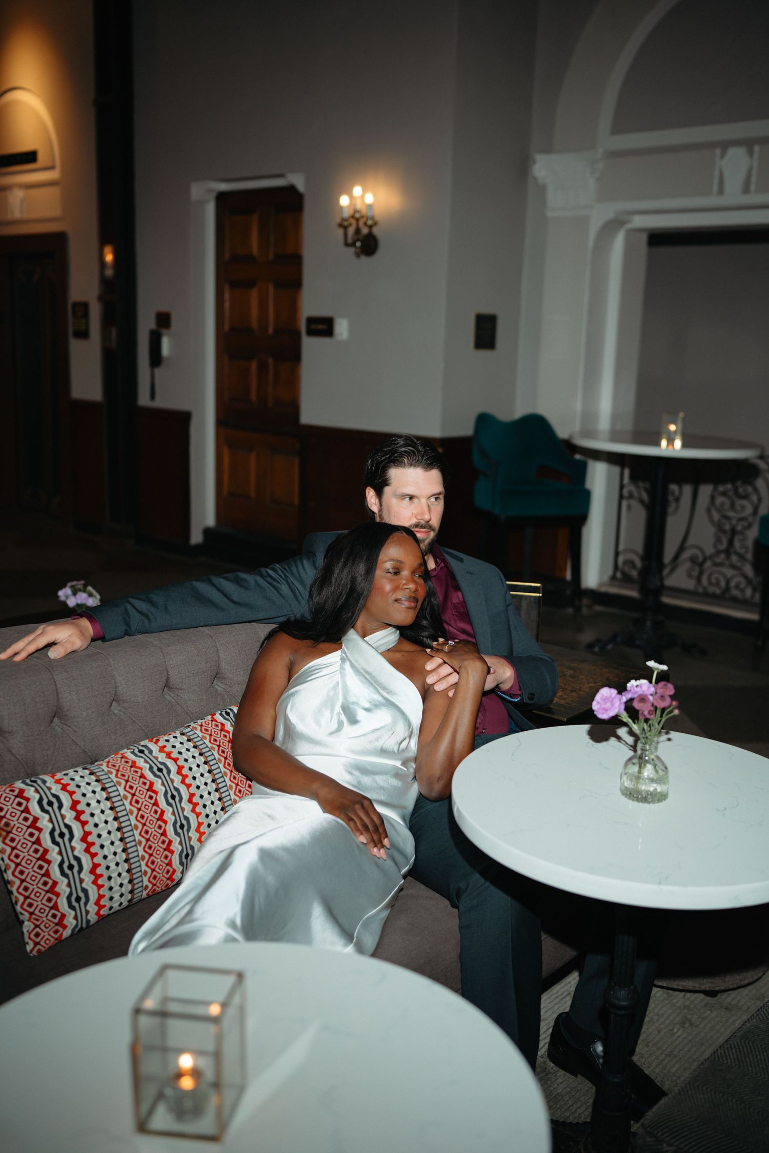 Interracial couple in wedding attire relaxing on a couch in an elegant dimly lit venue.