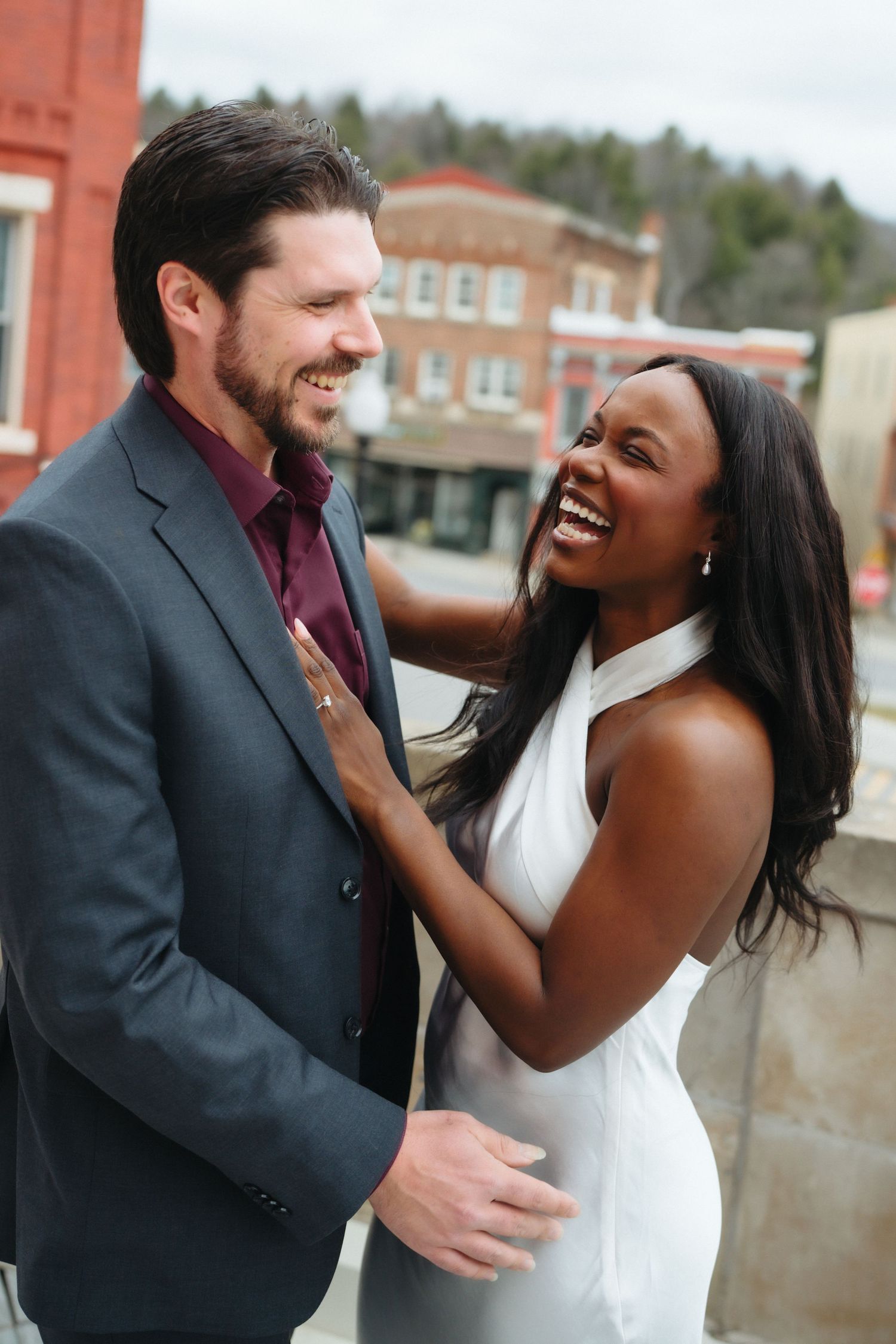 Joyful interracial couple laughing together outdoors, woman in white dress and man in gray suit, urban background.