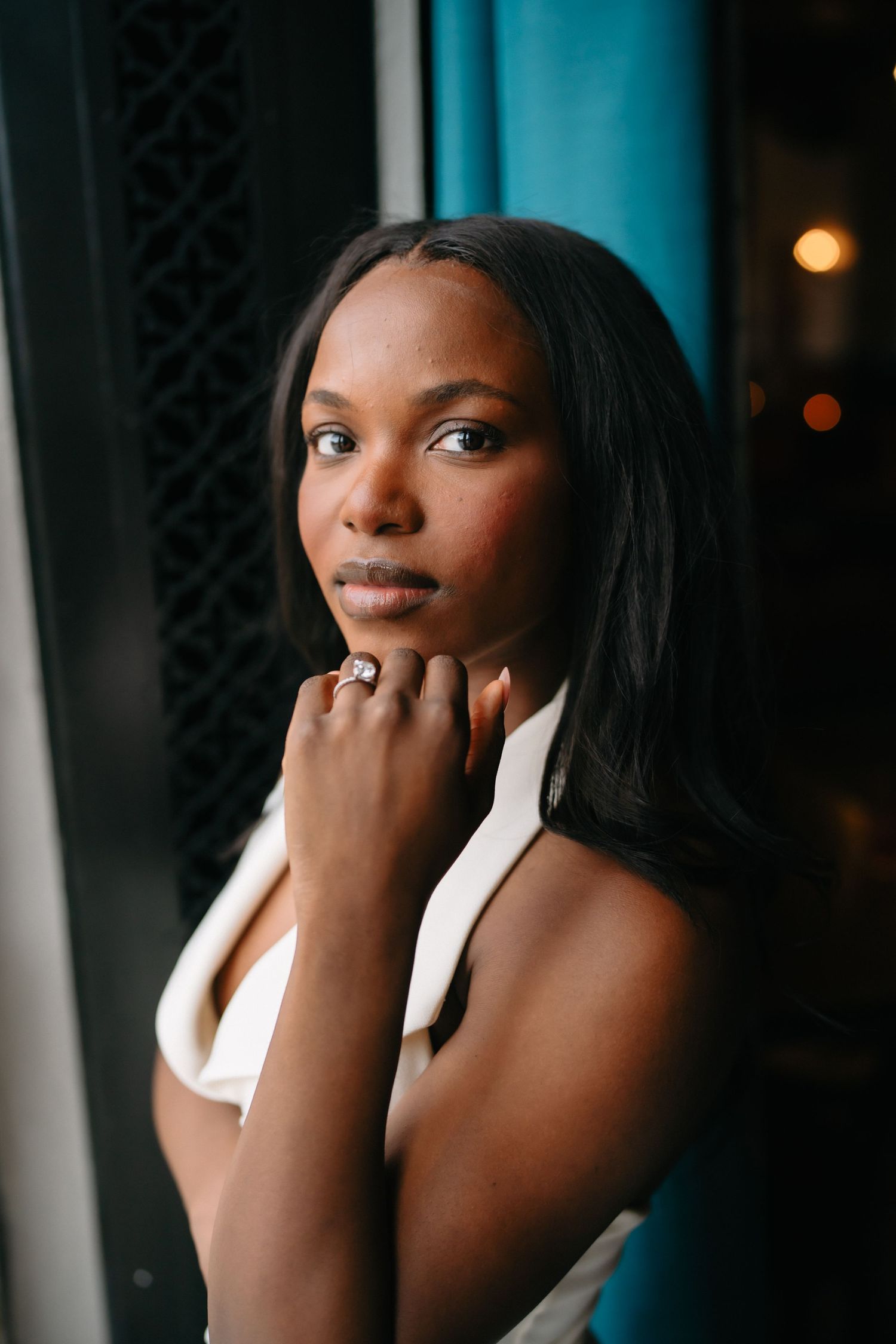 A young Black woman in a white halter top poses thoughtfully, hand on chin, wearing a ring, against a dark blue background.