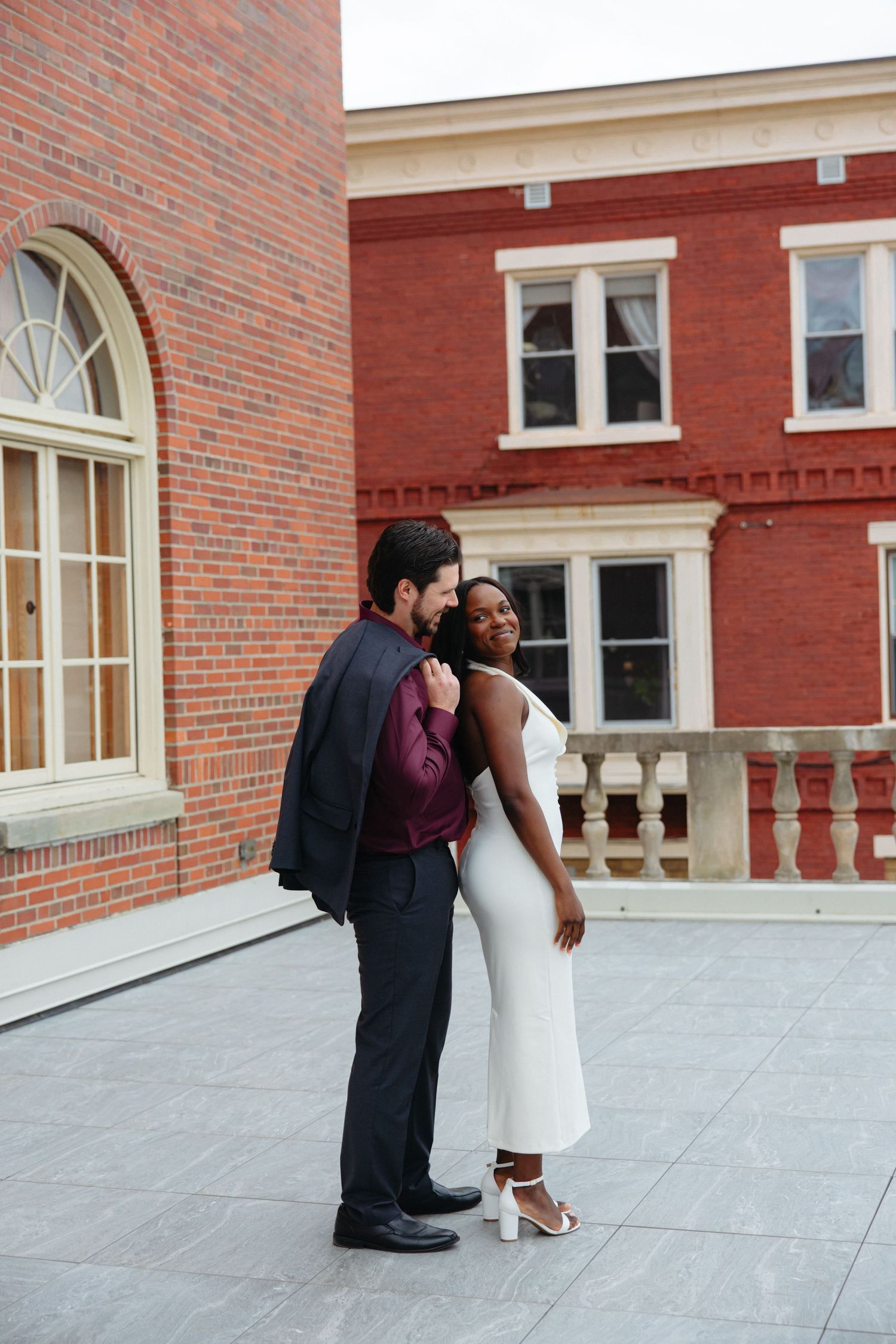 Interracial couple embracing on rooftop terrace of red brick building, woman in white dress and man in dark suit.