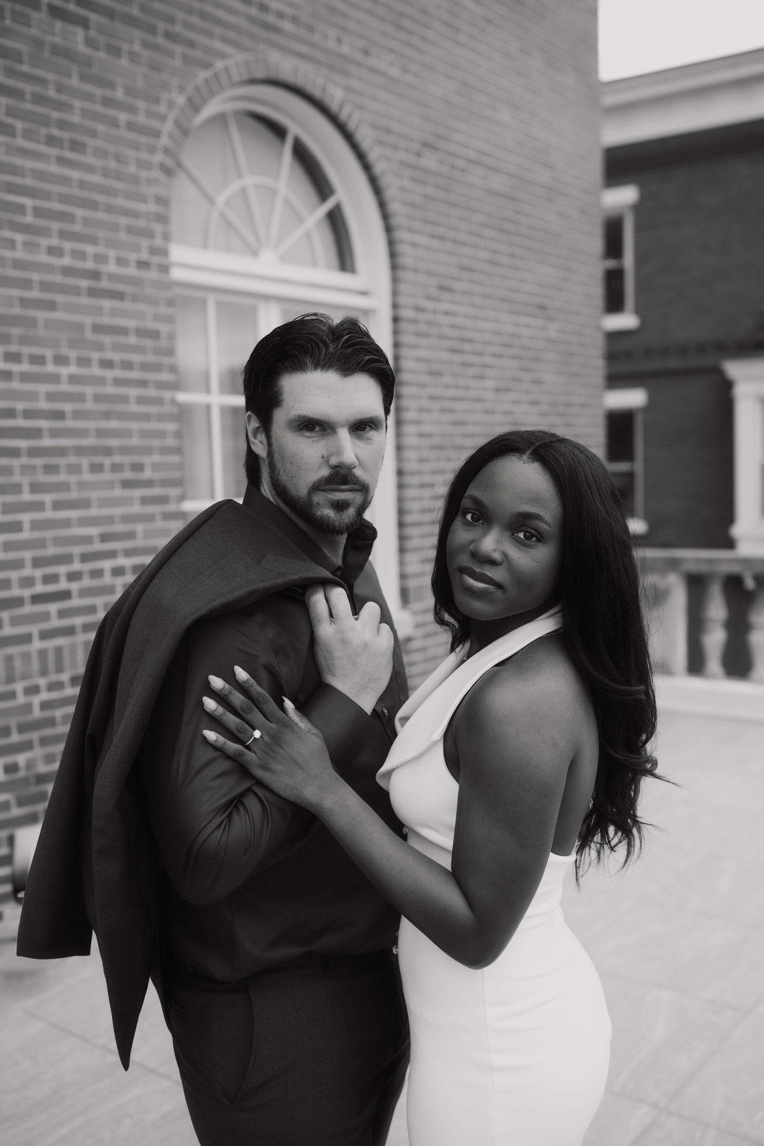 Black and white photo of an interracial couple posing elegantly outside a brick building, woman in white dress embracing suited man.