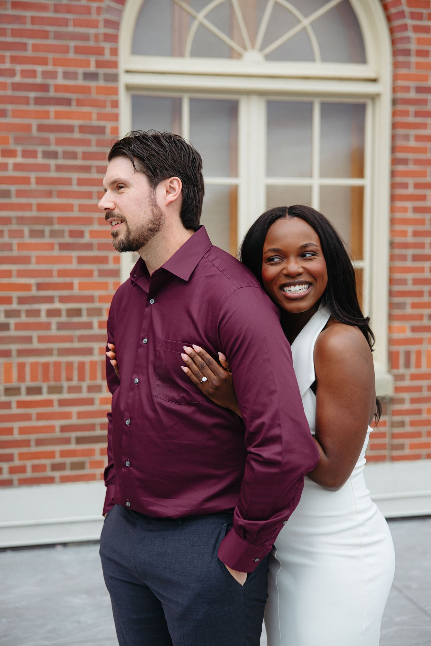 Interracial couple posing back-to-back, woman in white dress smiling, man in burgundy shirt, brick building backdrop.