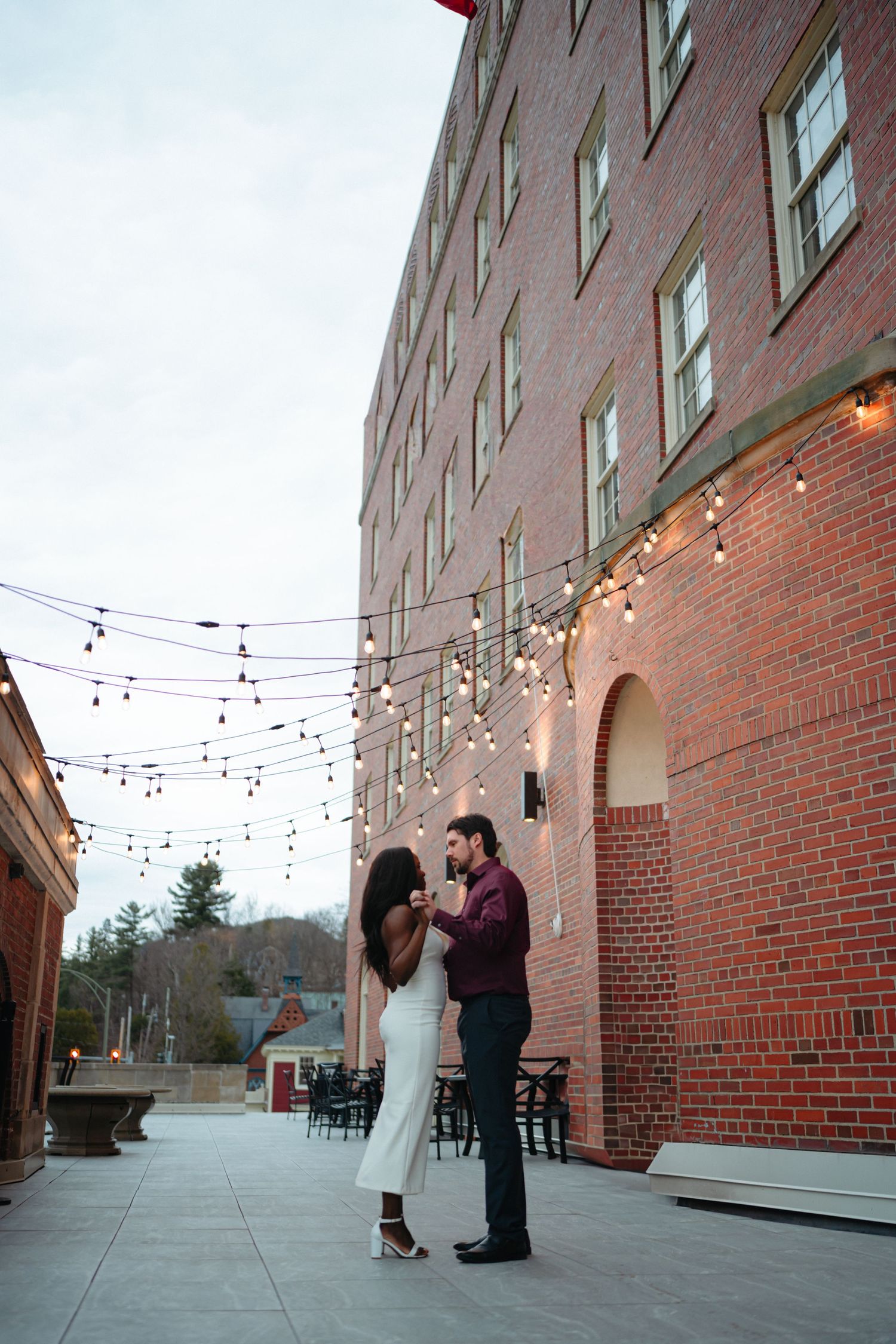 Couple dancing on a brick courtyard patio with string lights at dusk near a historic red brick building.