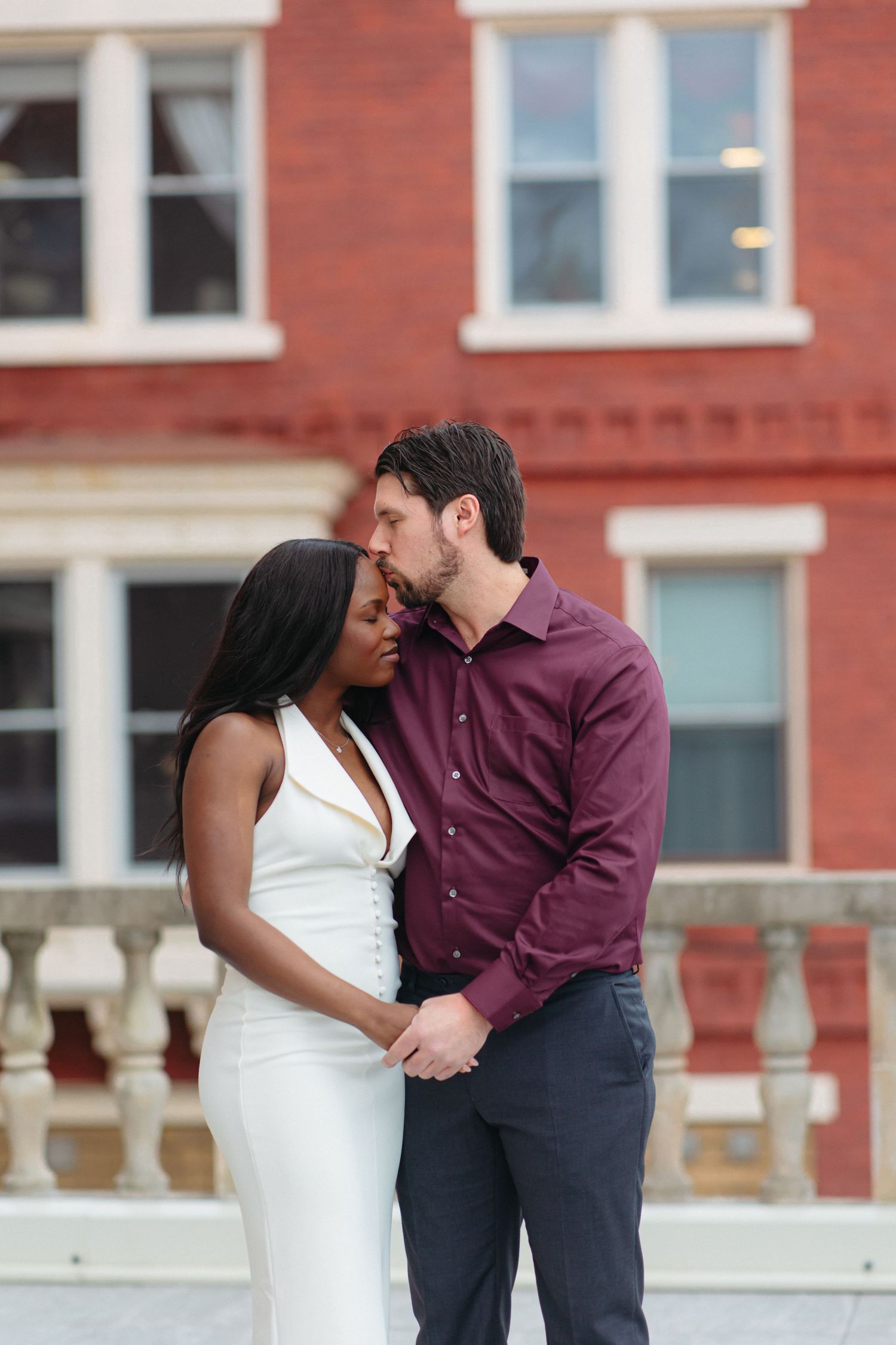 Interracial couple sharing a tender moment outdoors, man in burgundy shirt kissing woman in white dress near brick building.