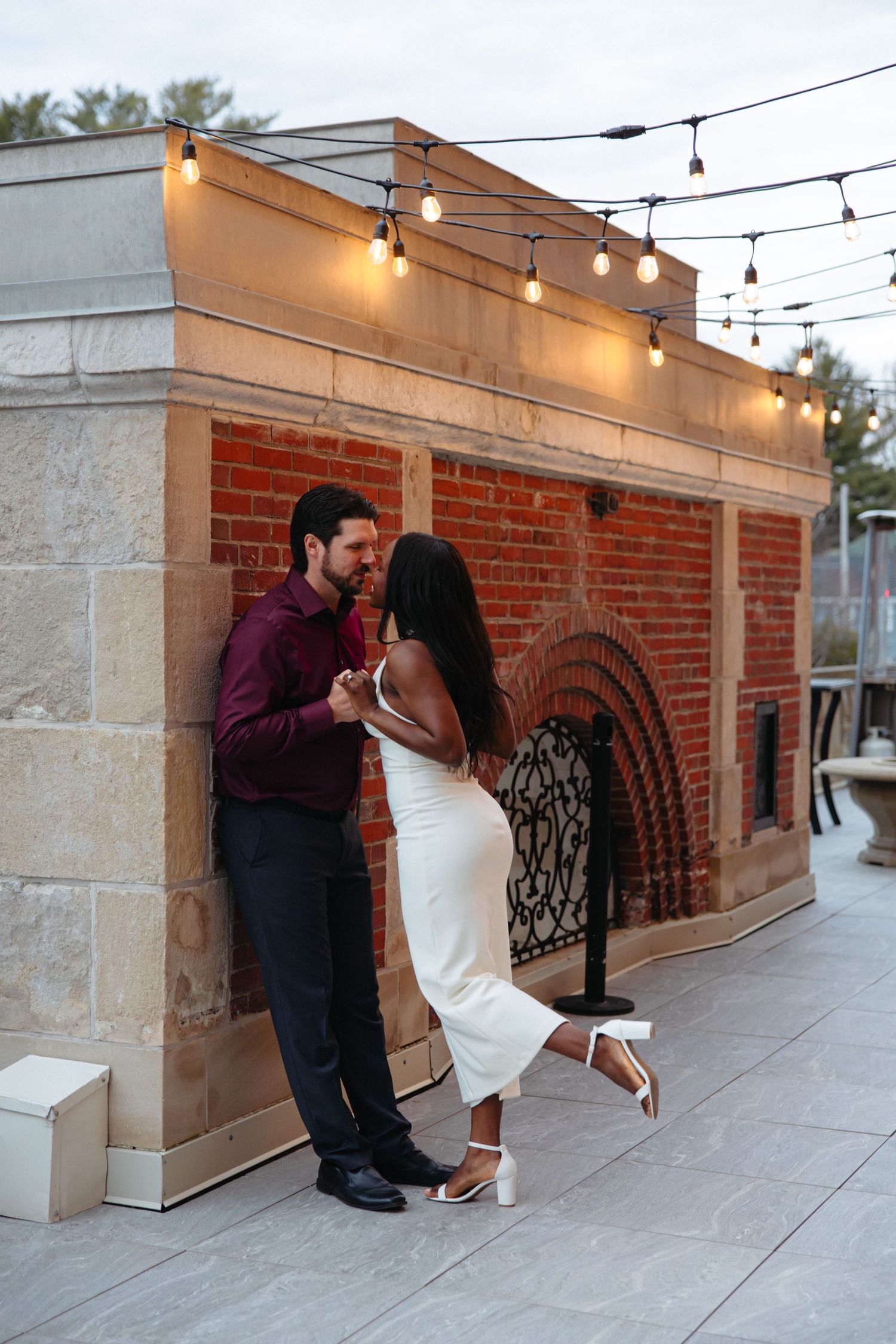 Romantic interracial couple sharing an intimate moment against a brick wall with string lights at dusk.