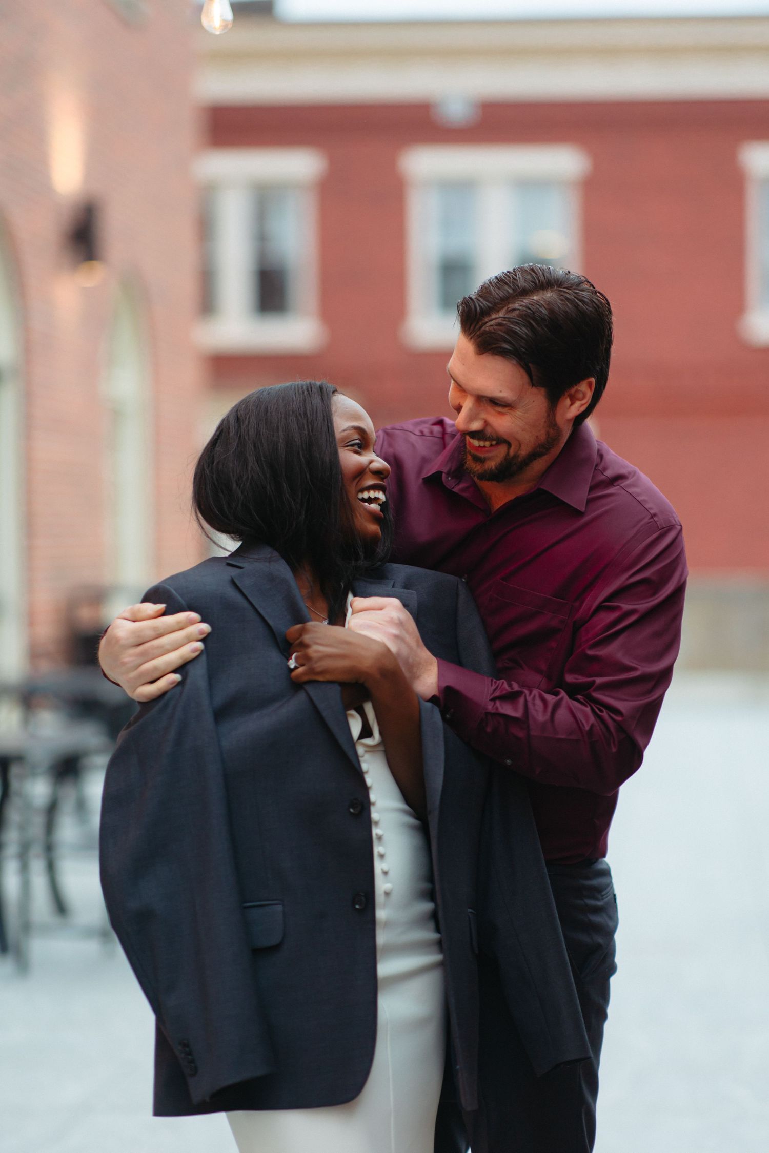 Man lovingly placing his jacket on a laughing woman outdoors near a red brick building.
