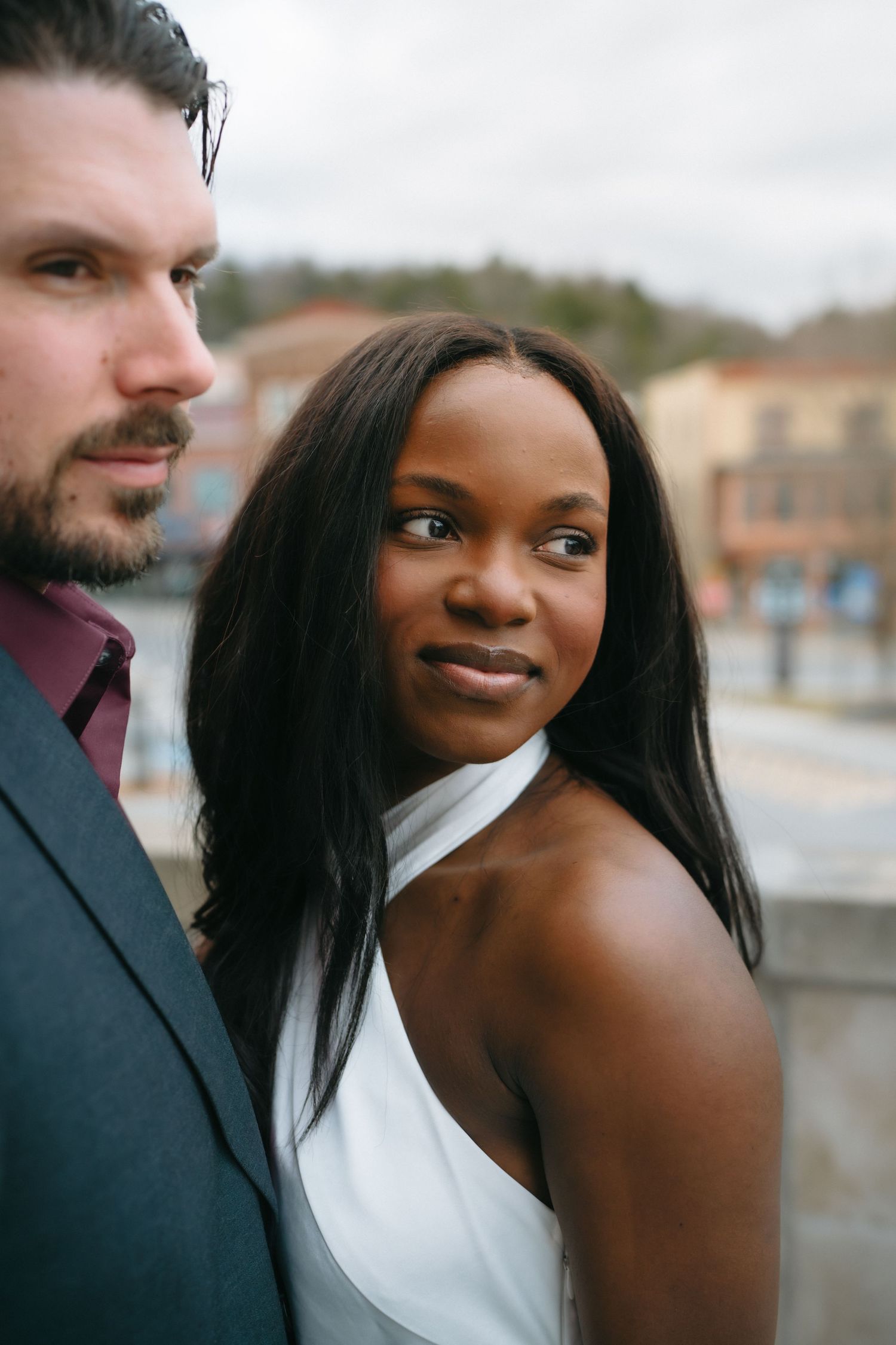 A Black bride in a white dress gazes back while standing with a bearded groom in a gray suit outdoors.