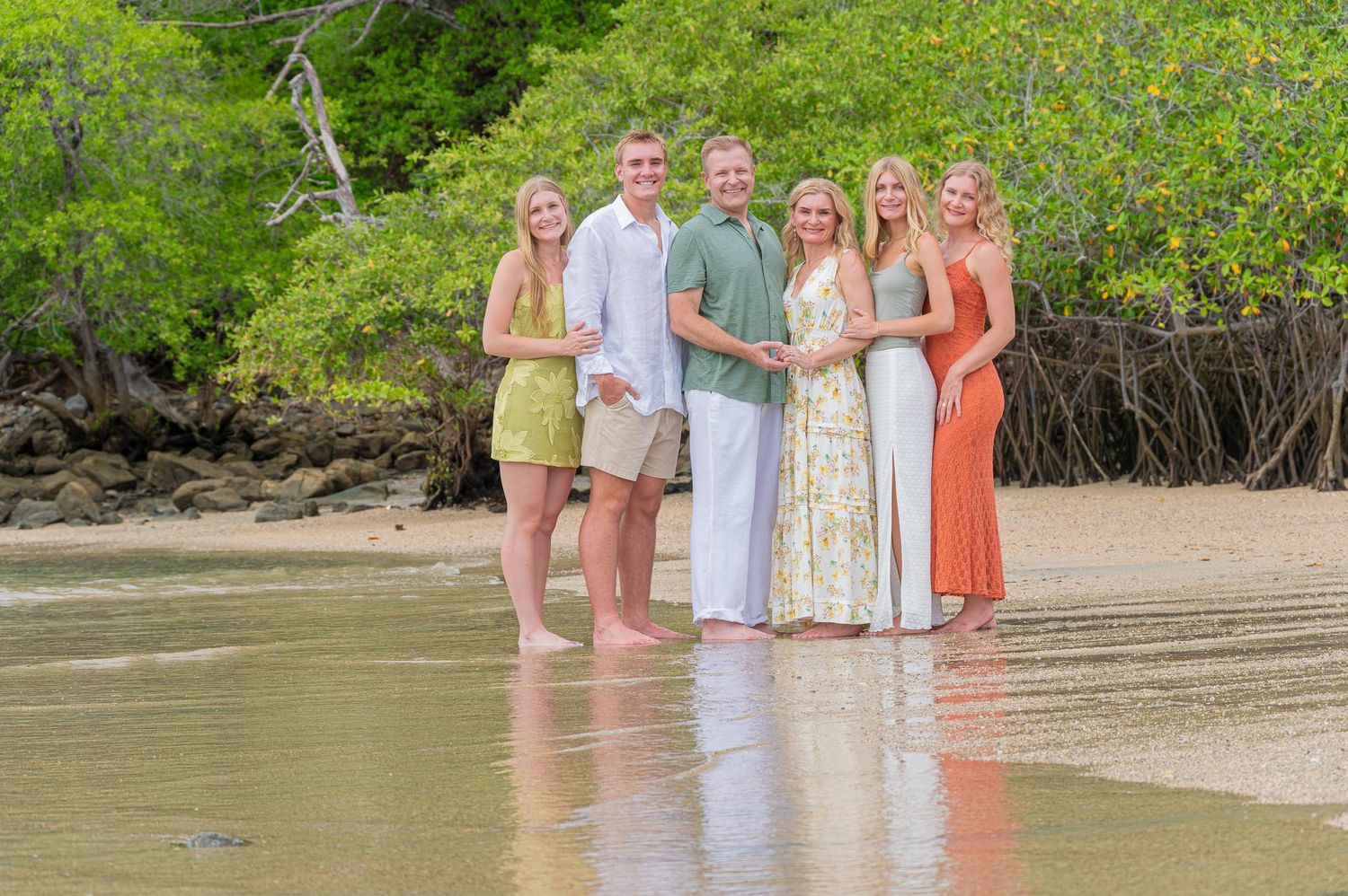 Family of six smiling together on a tropical beach with lush green mangroves in the background.