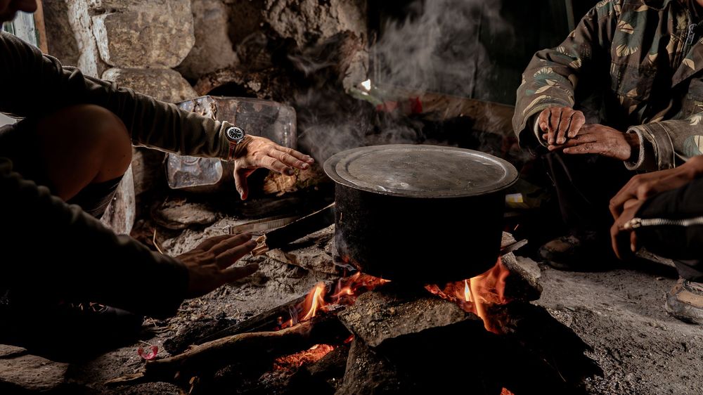 People cooking over an open fire with a large pot, warming hands in a dark rustic setting.