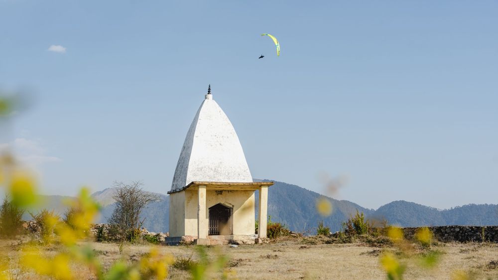 A white Hindu temple shrine stands on arid land with a paraglider soaring above against a clear blue sky and distant mountains.