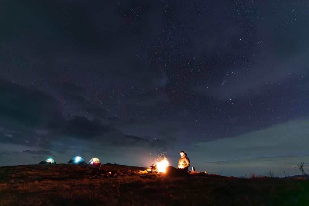 Person sitting by campfire under starry night sky with glowing tents on a dark hillside.
