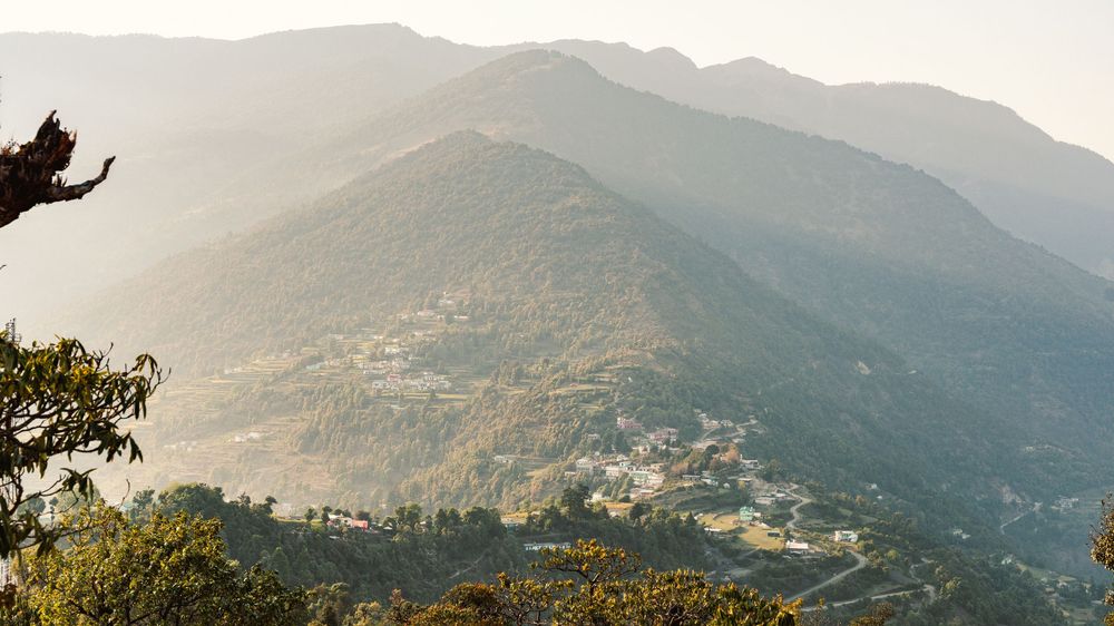 Aerial view of a mountain village nestled in lush green hills with misty peaks at golden hour.