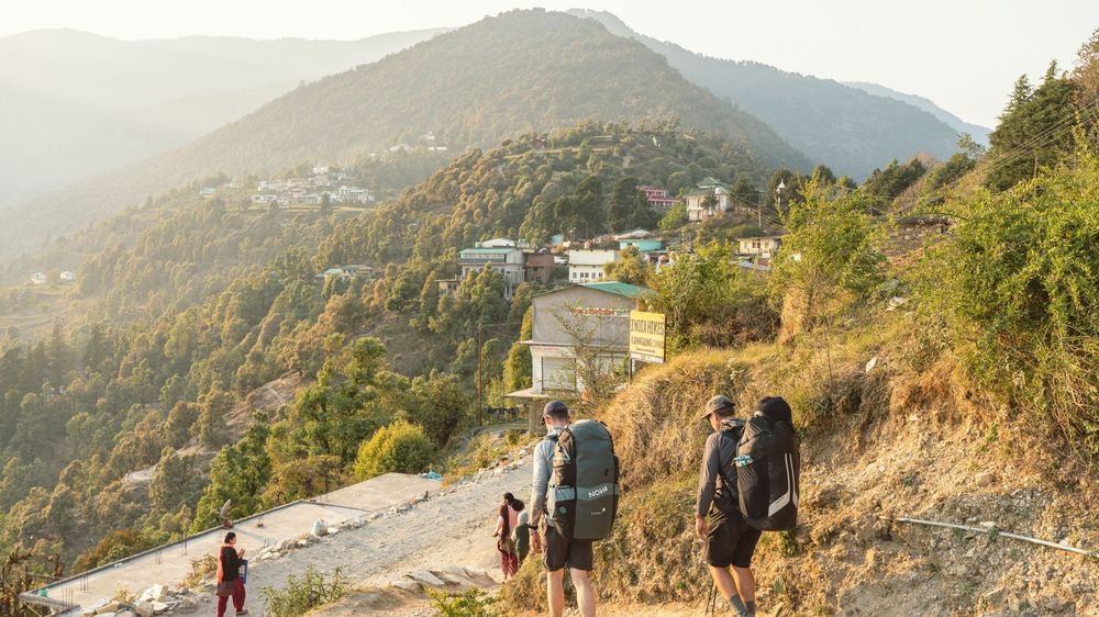 Hikers walking along a mountain road with forested hills and small village buildings in the golden sunset light.