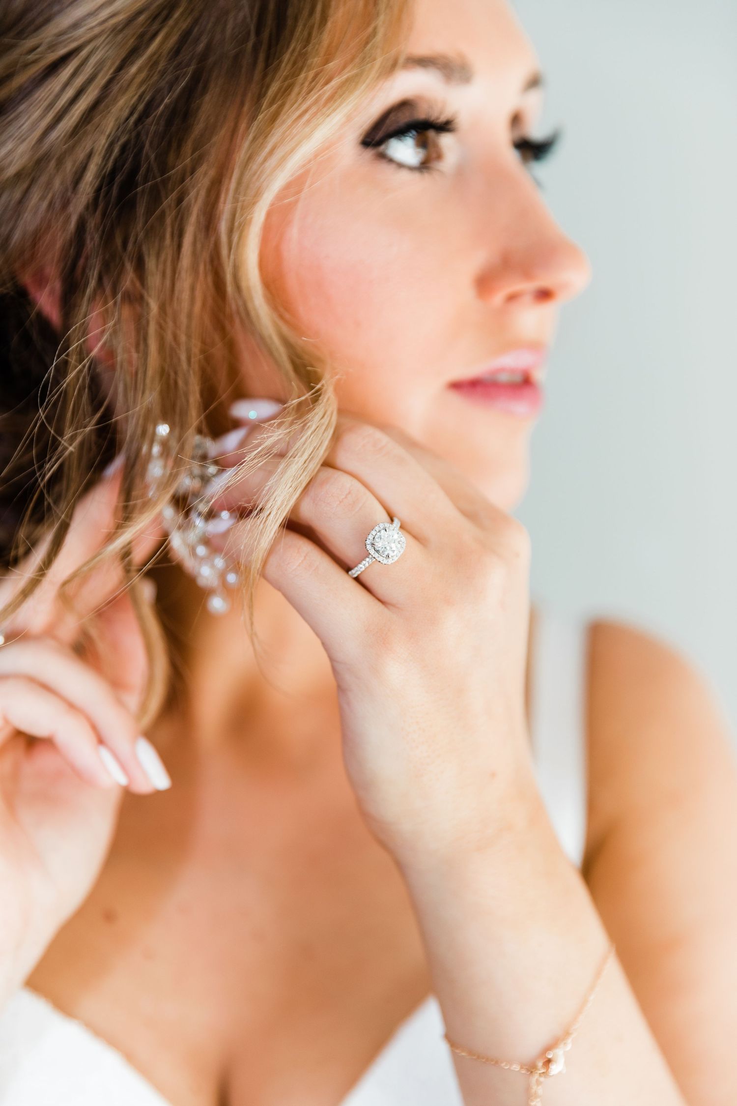 Bride adjusting pearl earring with diamond engagement ring visible, soft bridal makeup and updo hairstyle.