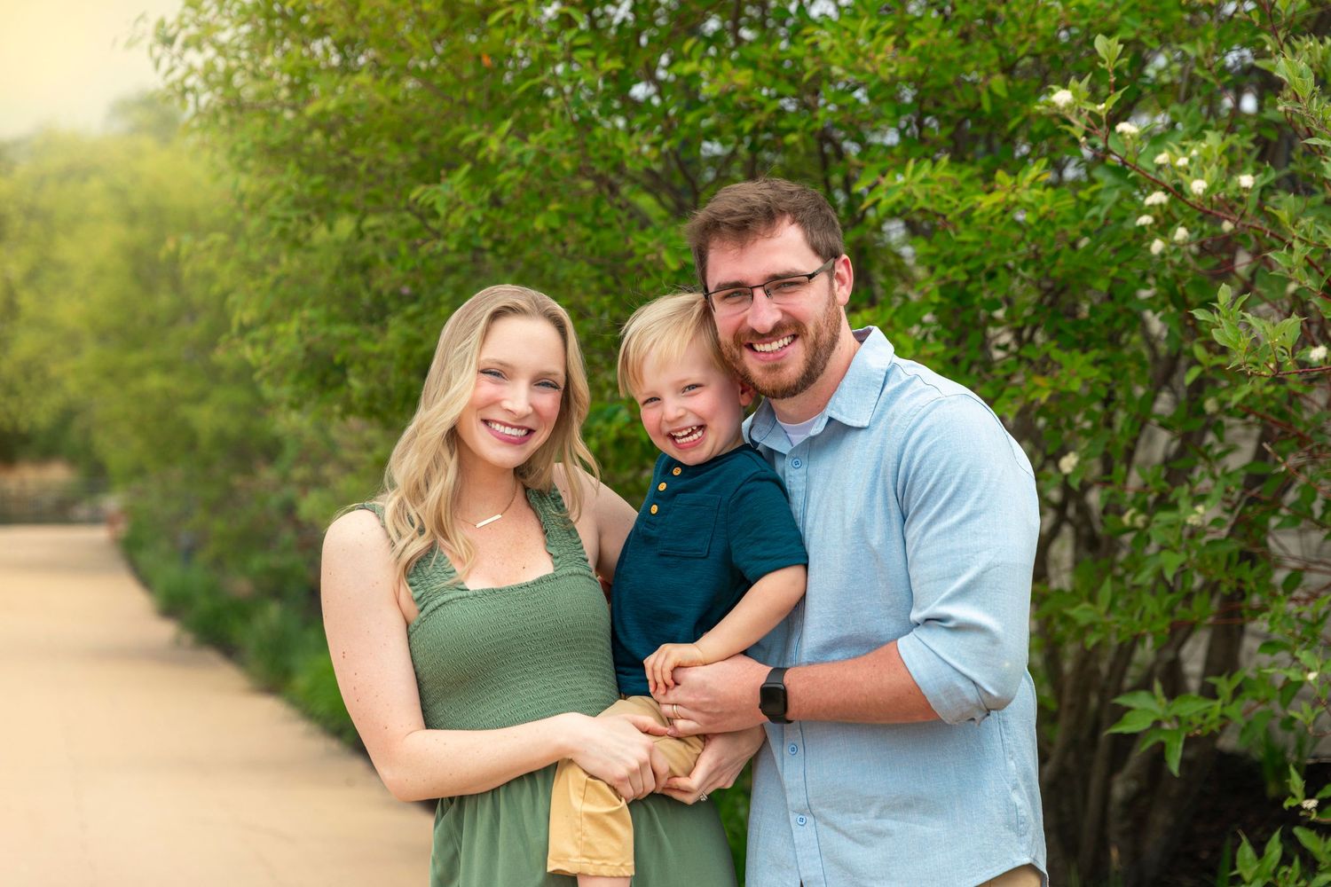 Happy family of three smiling outdoors, parents holding young boy, lush green trees in background.