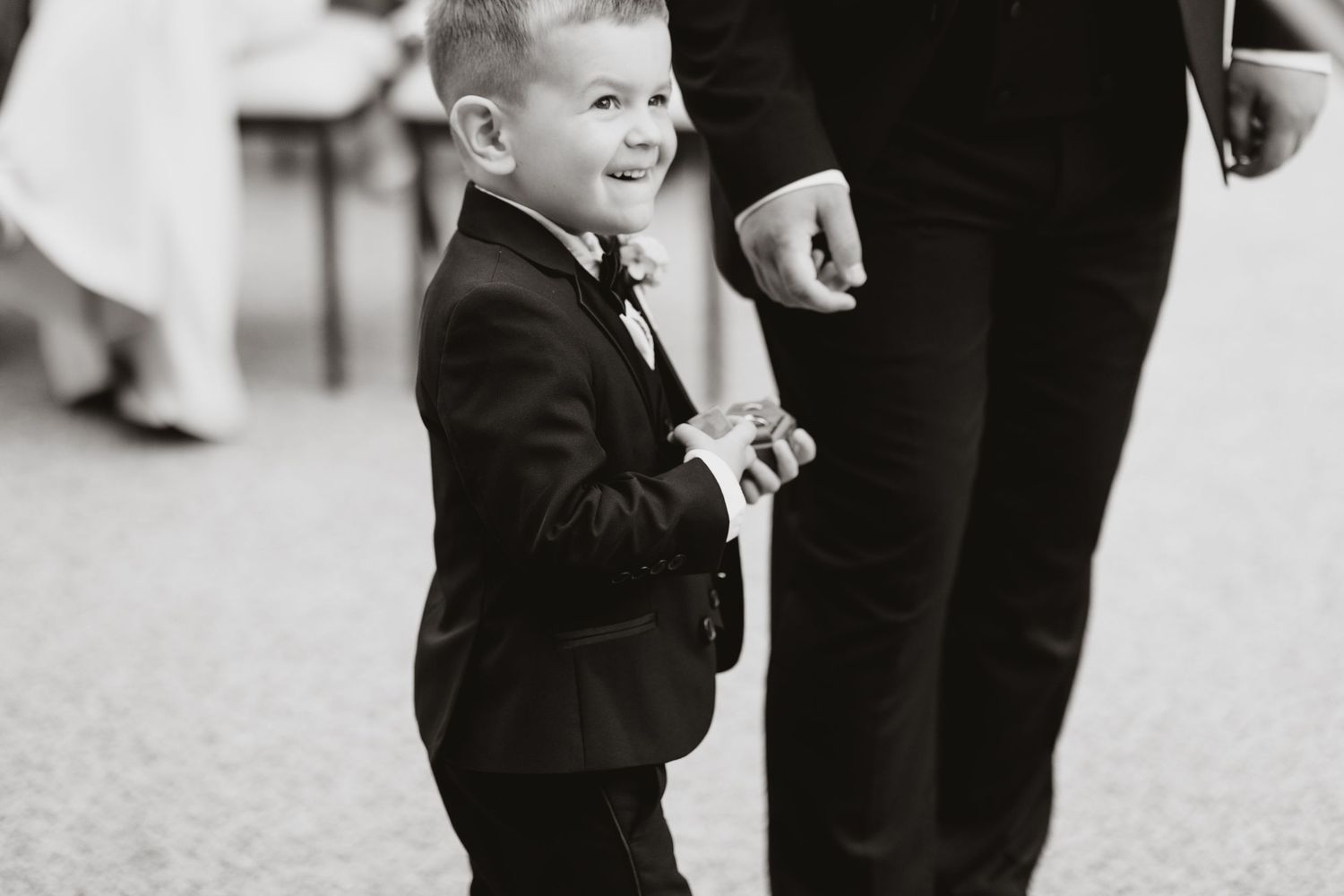 A joyful young boy in a black tuxedo and bow tie walks hand in hand with a man at a wedding.