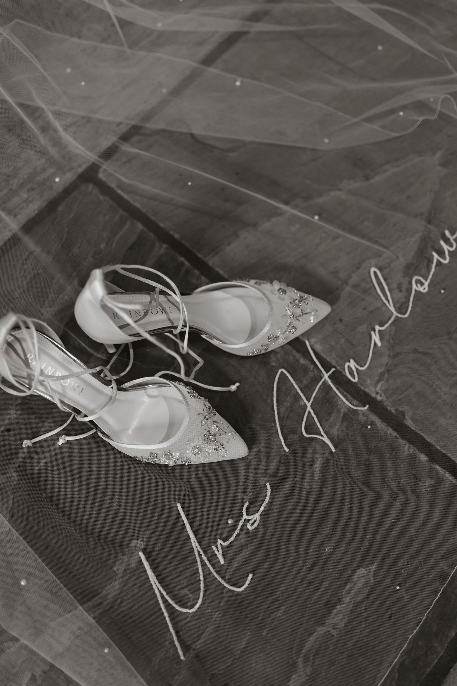 Black and white photo of embellished bridal heels on marble with a veil overlay and elegant cursive Miss Harlow script.