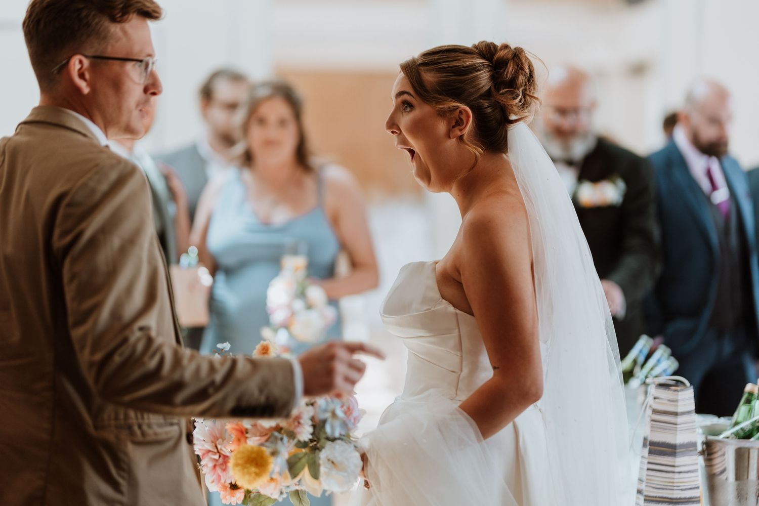 Bride in white strapless gown and veil laughing joyfully during wedding ceremony with guests in background.