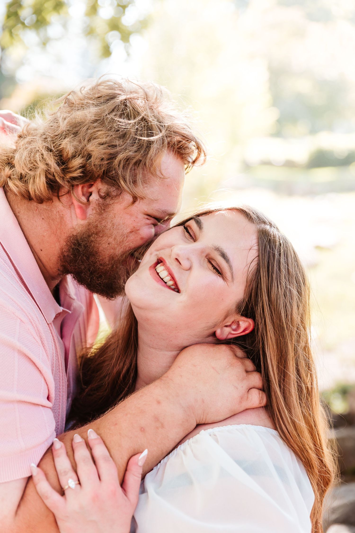 A bearded man with curly hair kisses a laughing woman outdoors in warm sunlight.