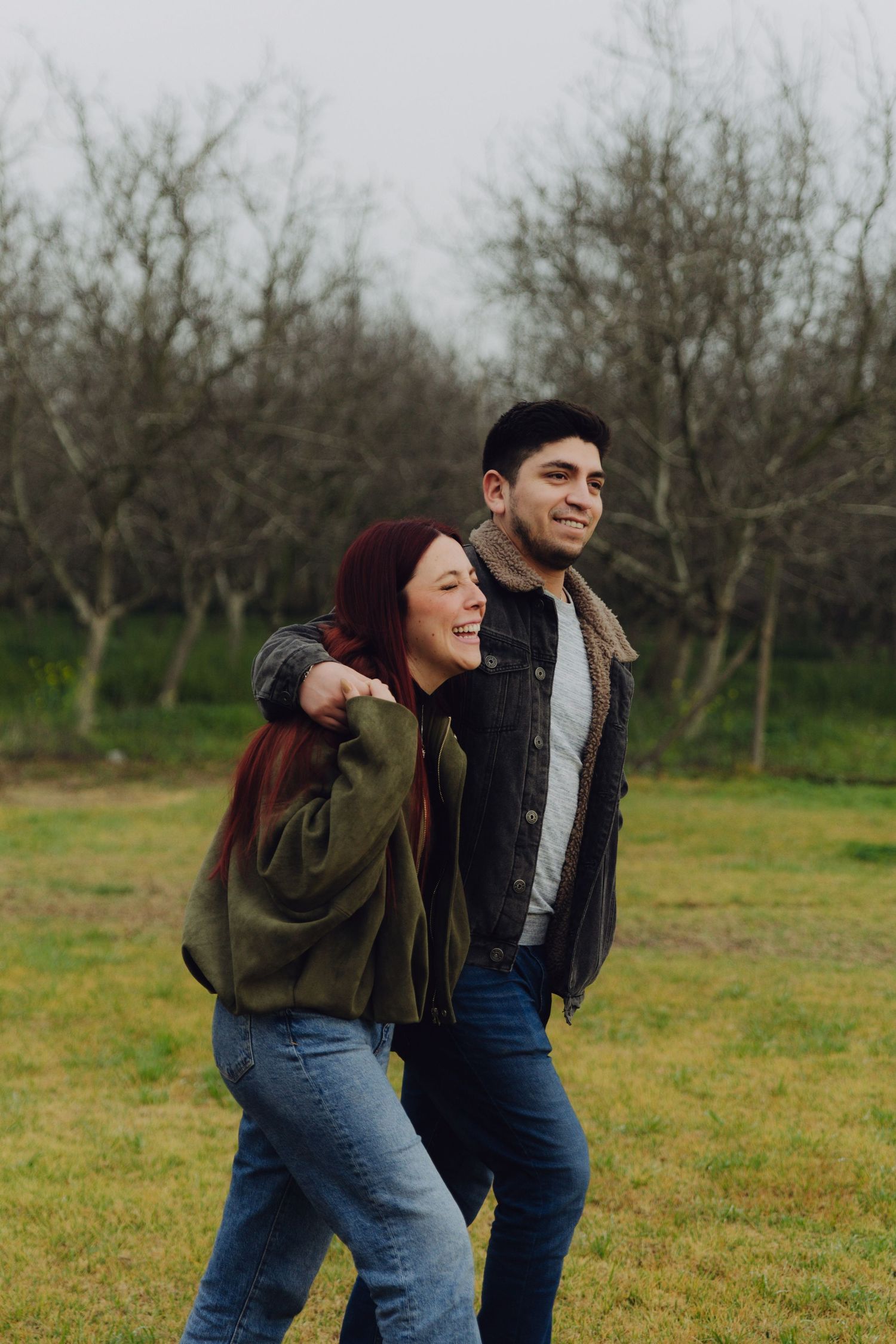 A laughing couple walks playfully through a grassy field with bare winter trees in the background.