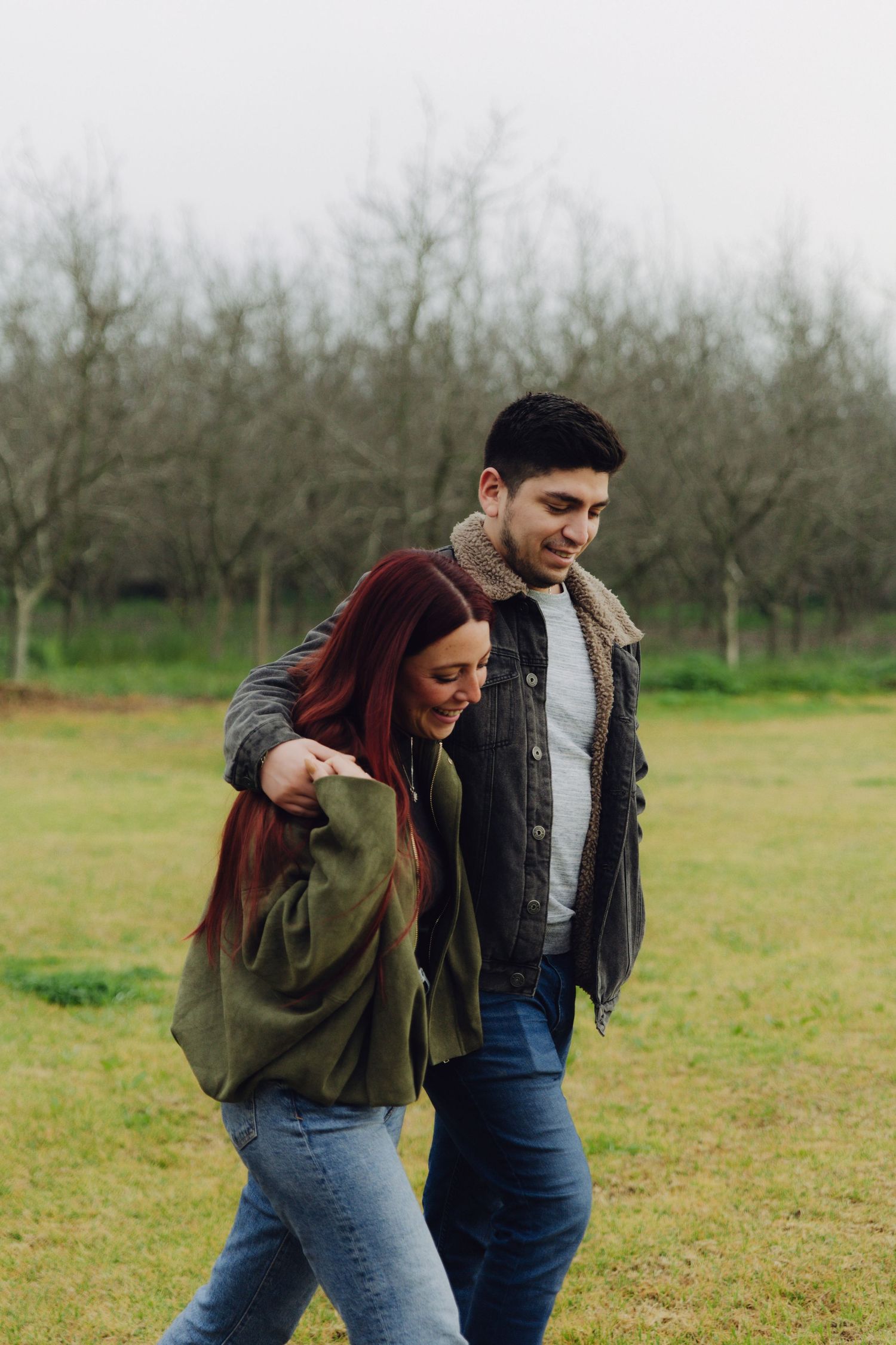 A young couple walks arm in arm across a green field surrounded by bare trees on an overcast day.