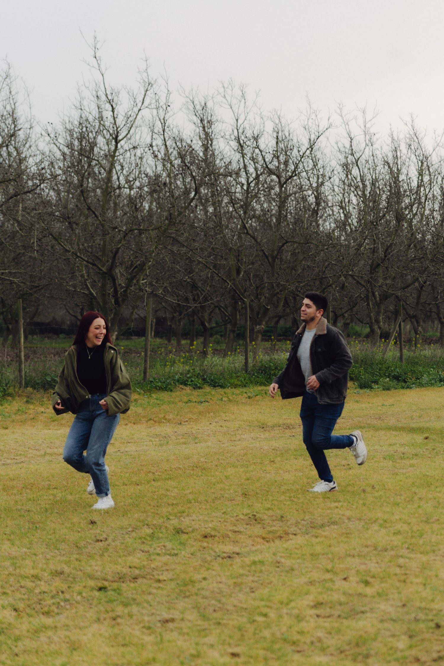 A joyful couple runs and plays together across a grassy field with bare trees in the background.