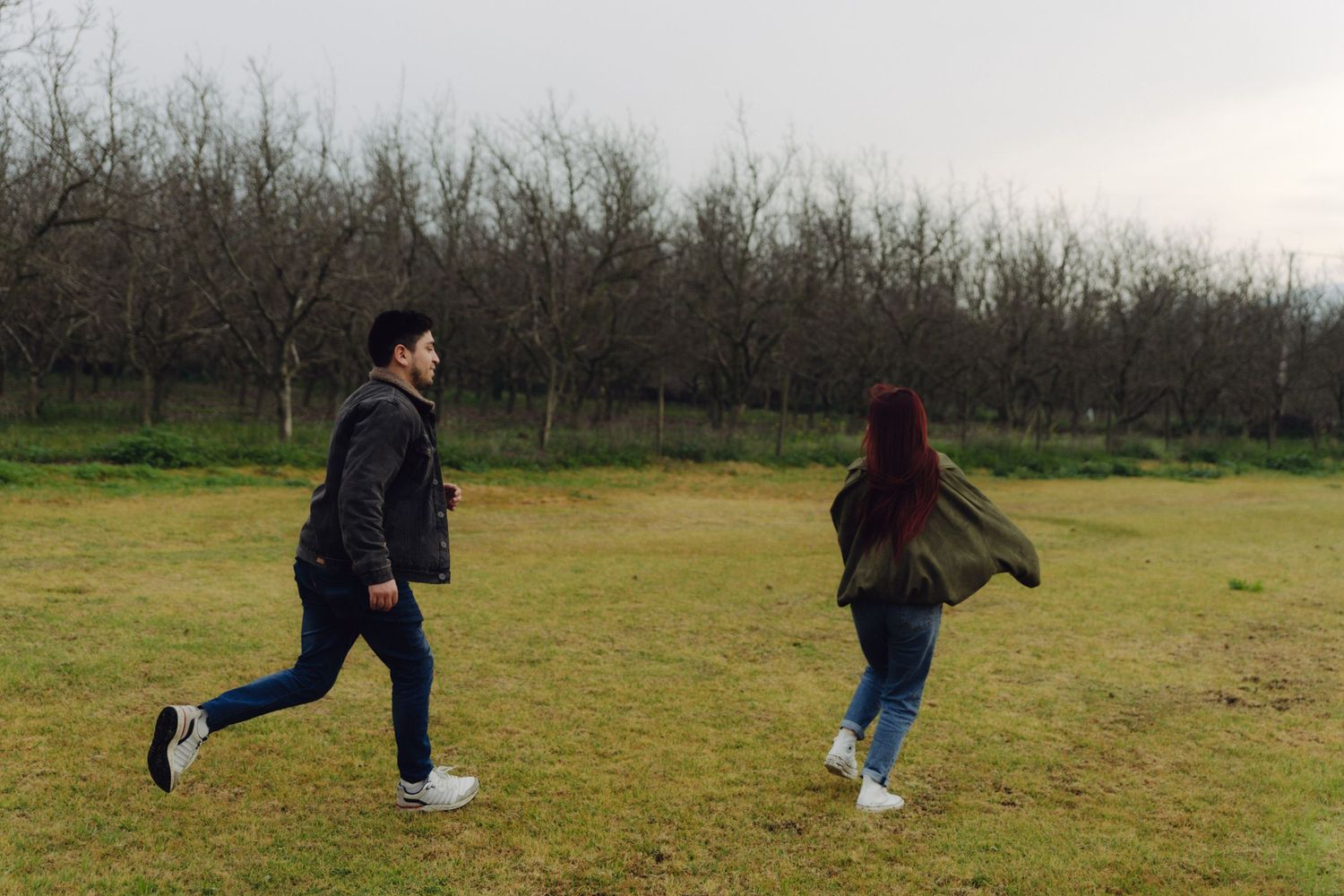 A couple runs playfully across a green field with rows of bare winter trees in the background.