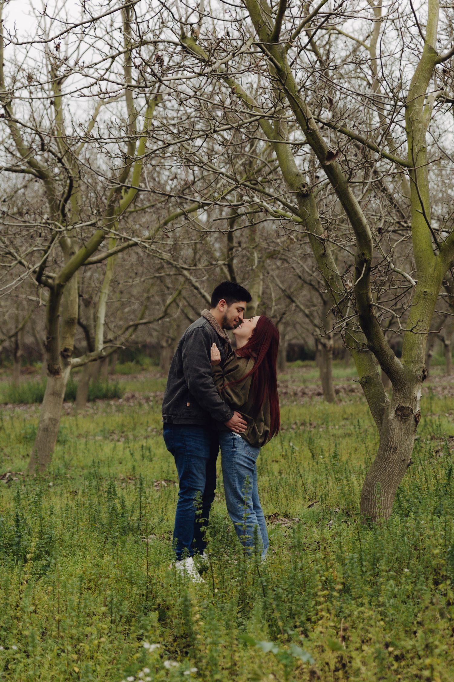A couple shares a romantic kiss among bare trees in a green meadow on an overcast day.
