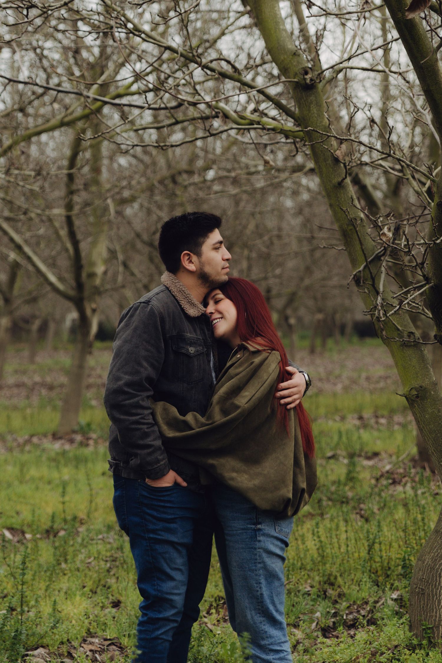 Couple embracing in a bare winter orchard, woman with red hair smiling against man in dark jacket.