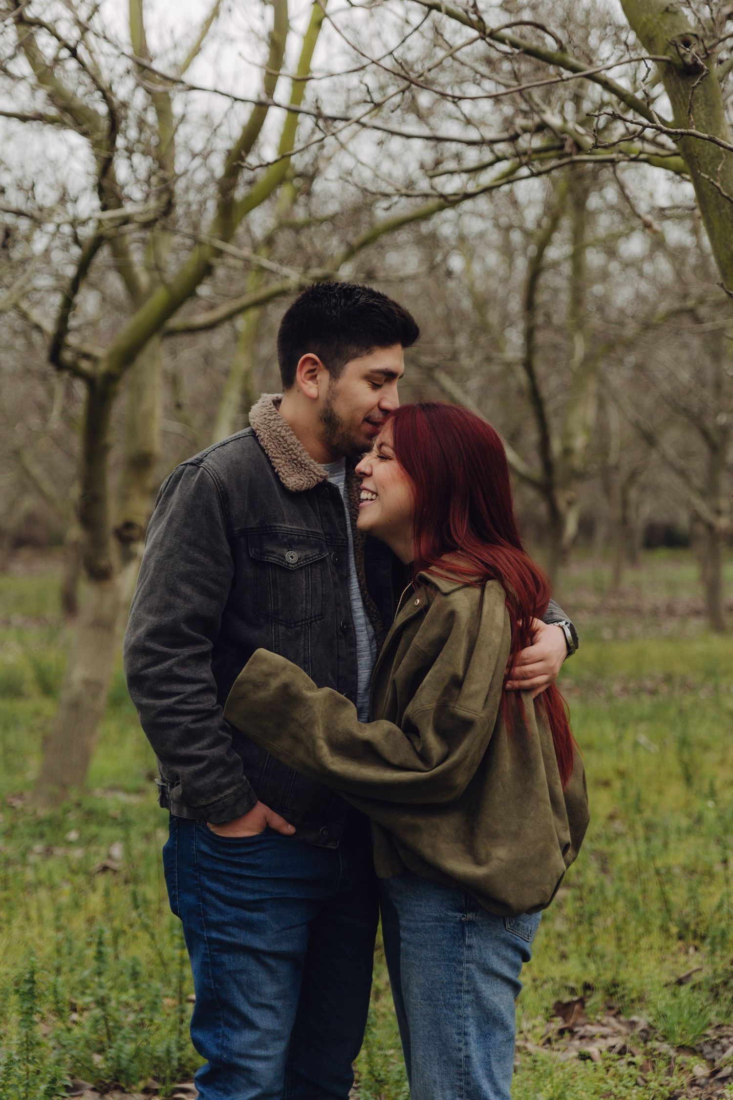 Smiling couple embracing tenderly among bare trees in a green grassy woodland setting.