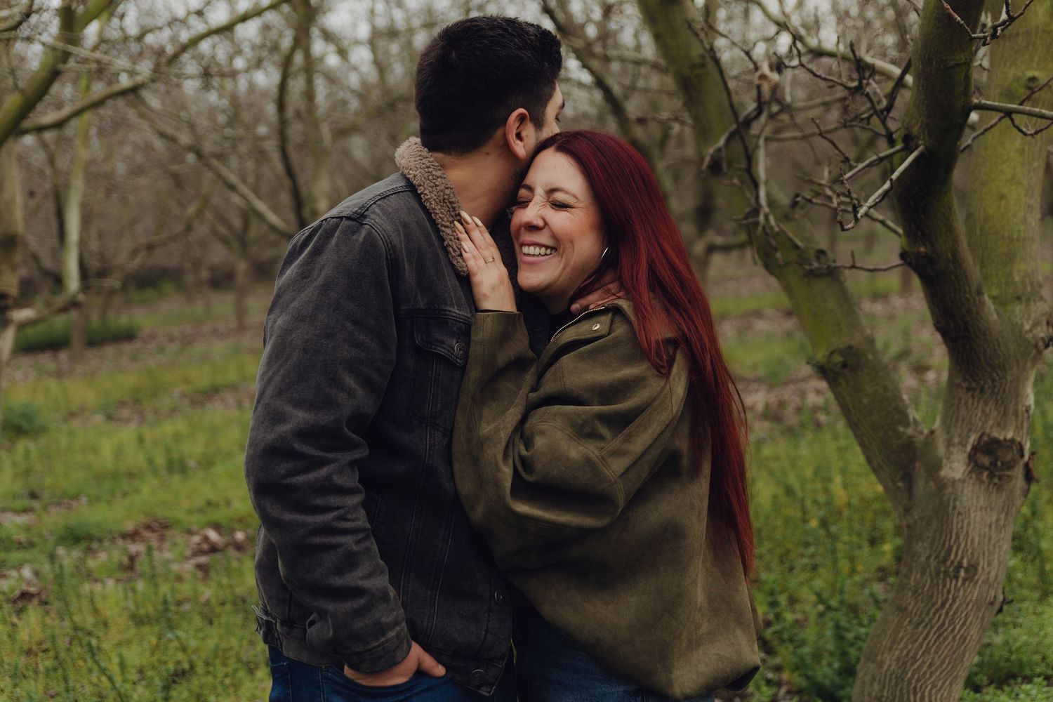 Red-haired woman laughing joyfully as her partner whispers to her in a bare winter orchard.