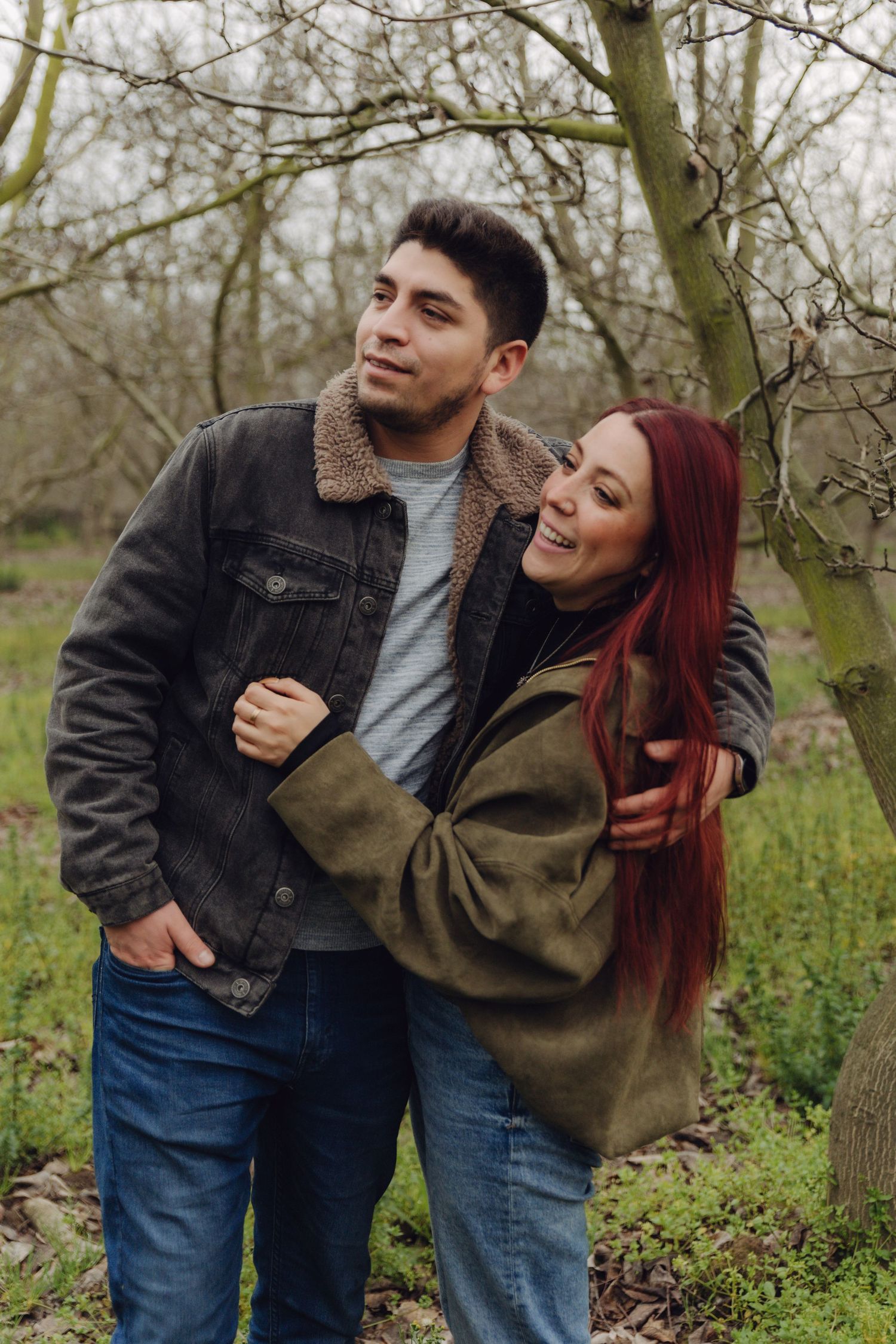 Happy couple embracing outdoors in a bare winter orchard, woman with red hair smiling brightly.