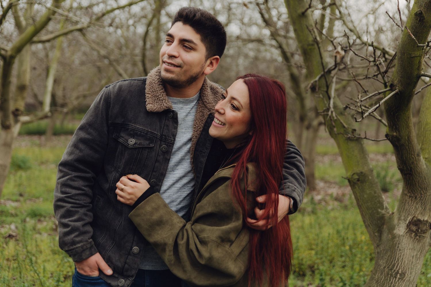 Cheerful couple holding each other among bare trees in a lush green outdoor setting.