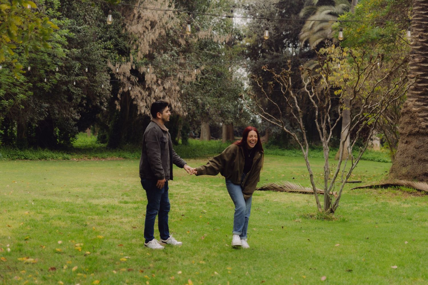 A couple holds hands and laughs playfully on a green lawn surrounded by trees in a park.