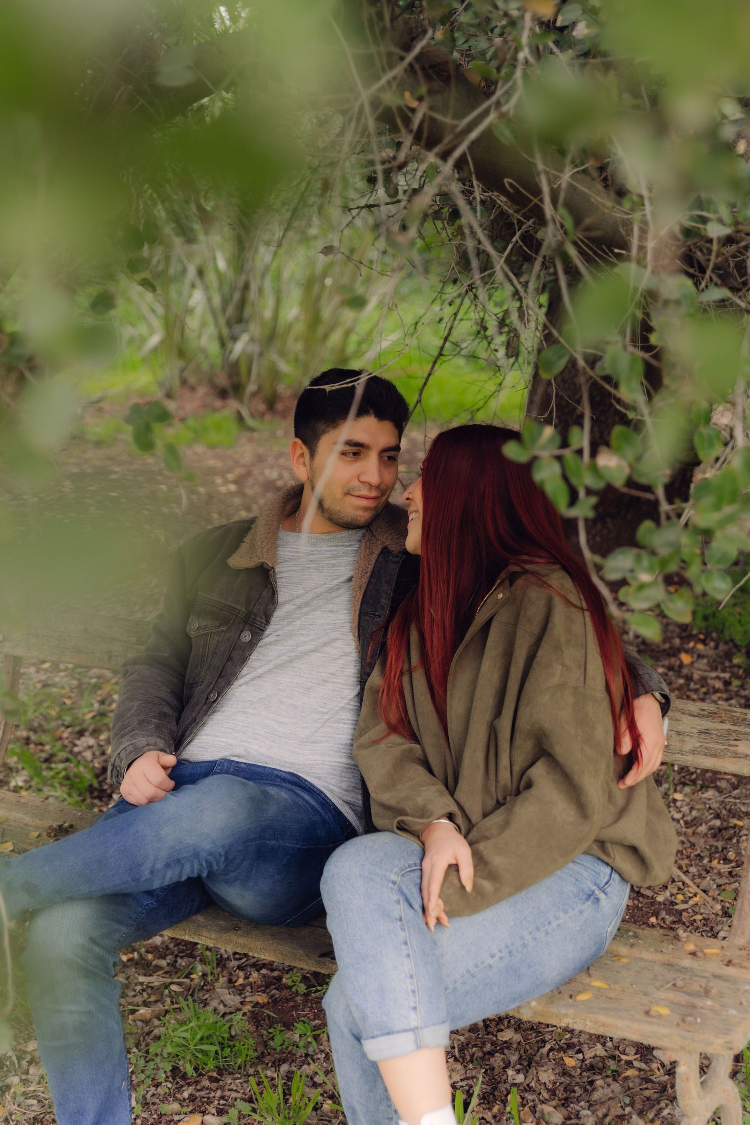 A young couple sits closely together on a bench surrounded by lush greenery in a park.