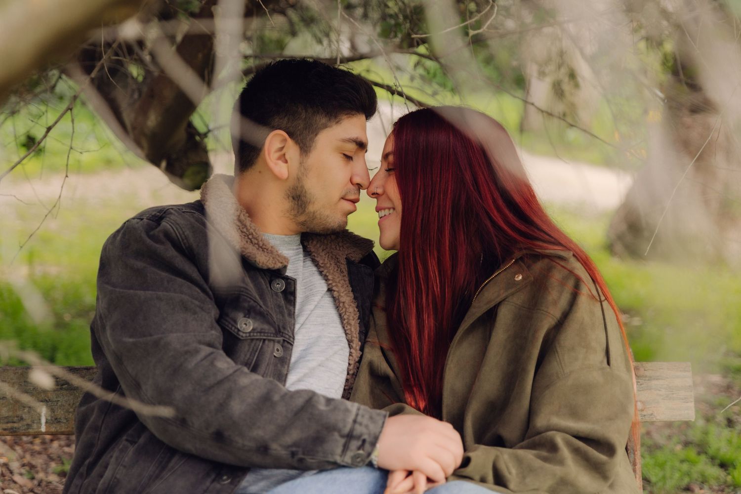 A romantic couple leans in for a kiss while sitting under a tree in a park.