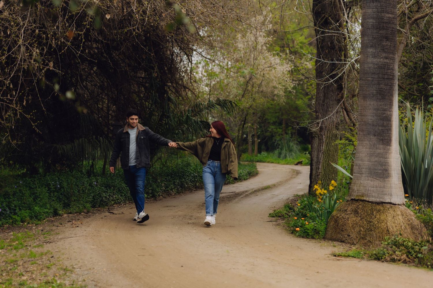 A couple walks hand in hand down a winding dirt path lined with trees and flowers.