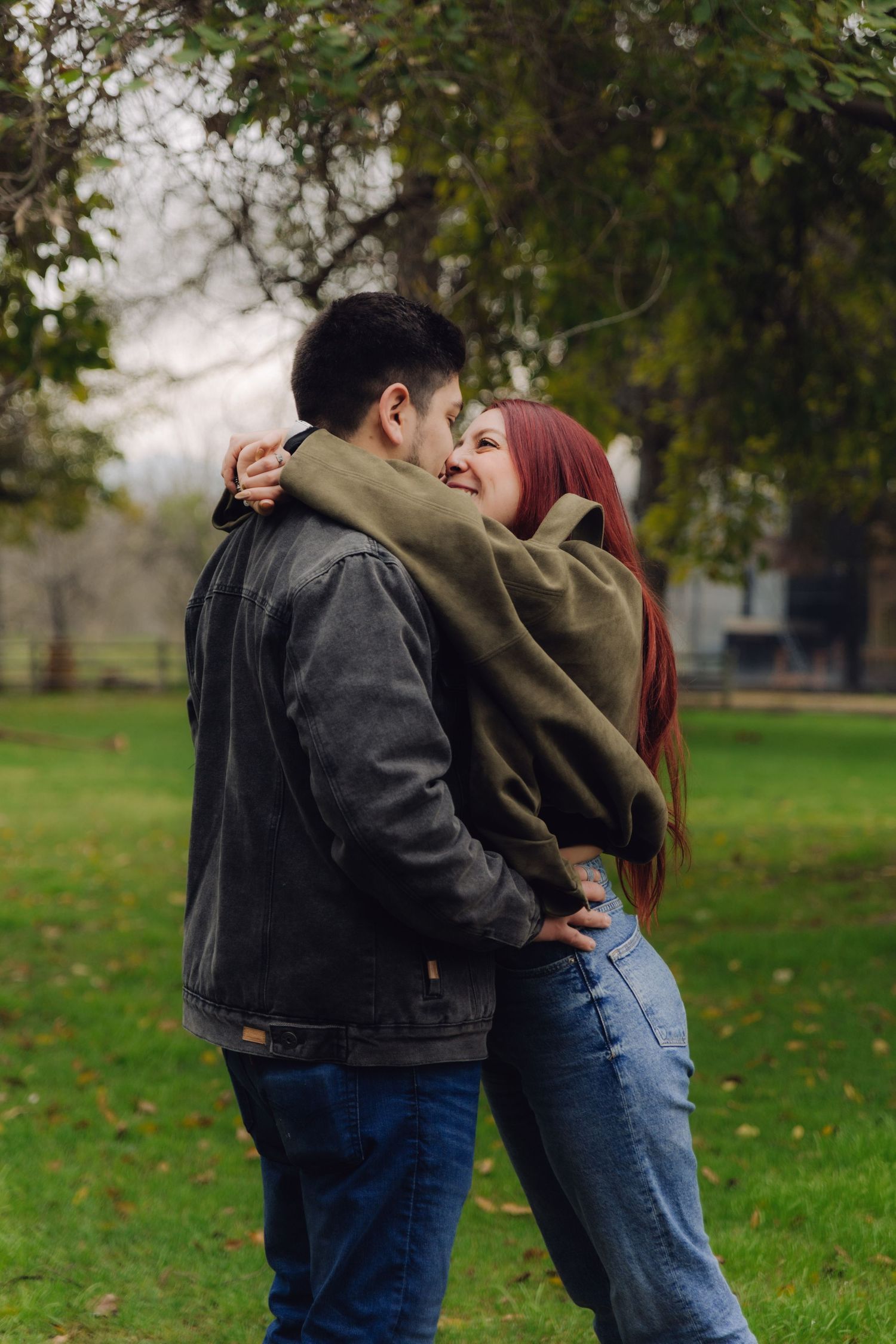 A laughing red-haired woman hugs a man in a denim jacket on a green lawn.