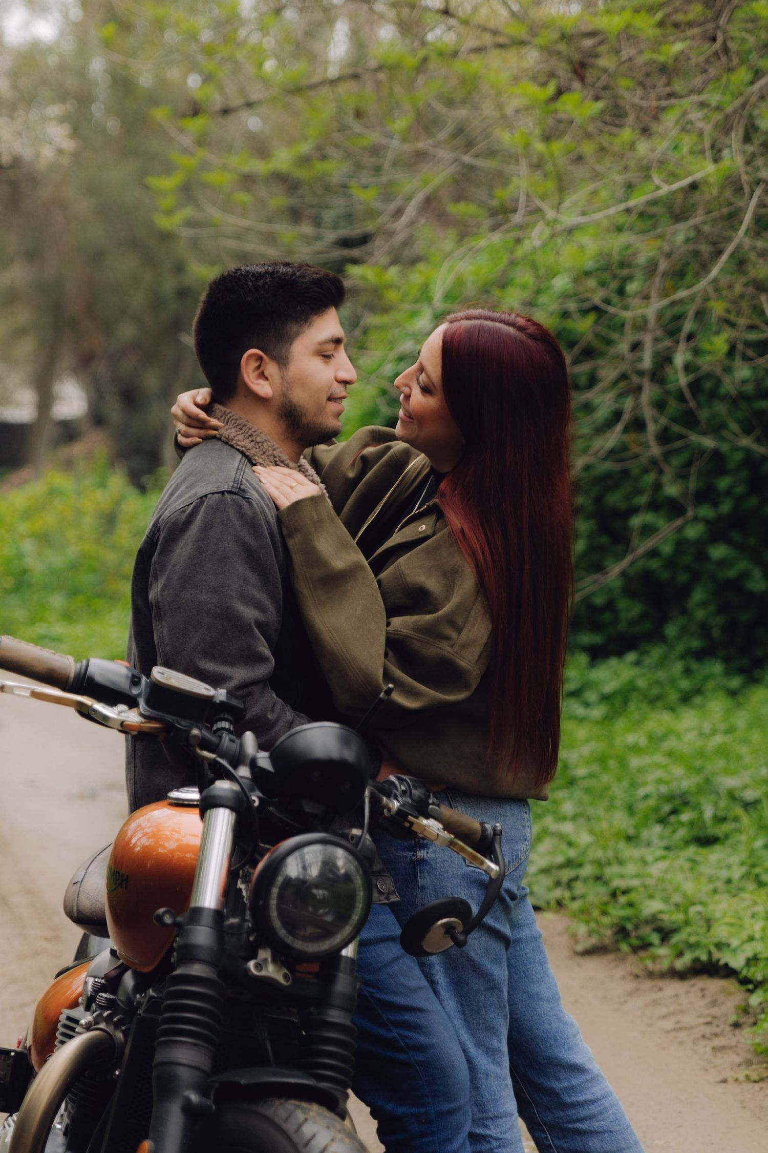 A romantic couple embraces near a vintage motorcycle on a tree-lined outdoor path.