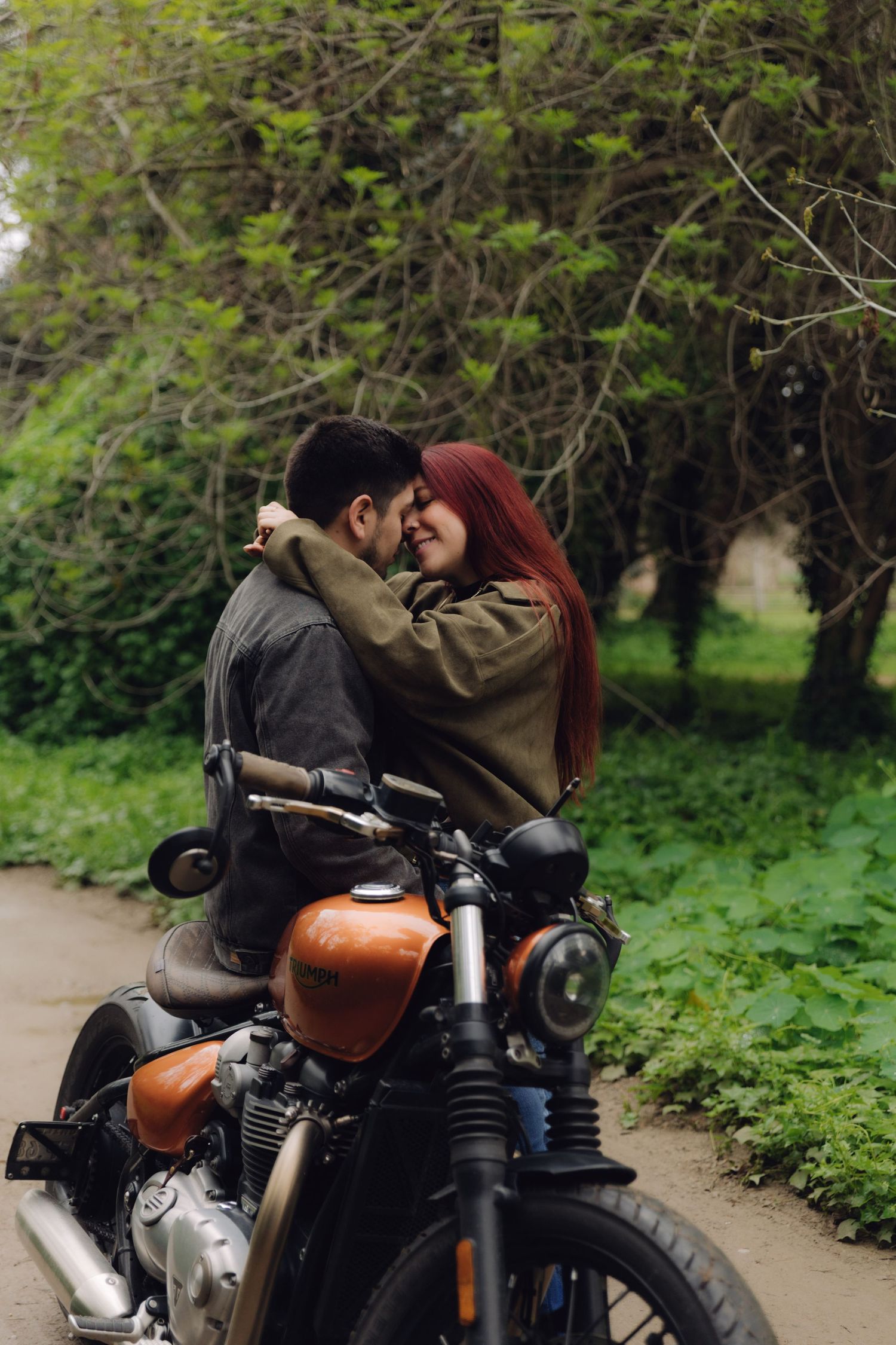 Romantic couple embracing on a vintage orange motorcycle on a tree-lined path.