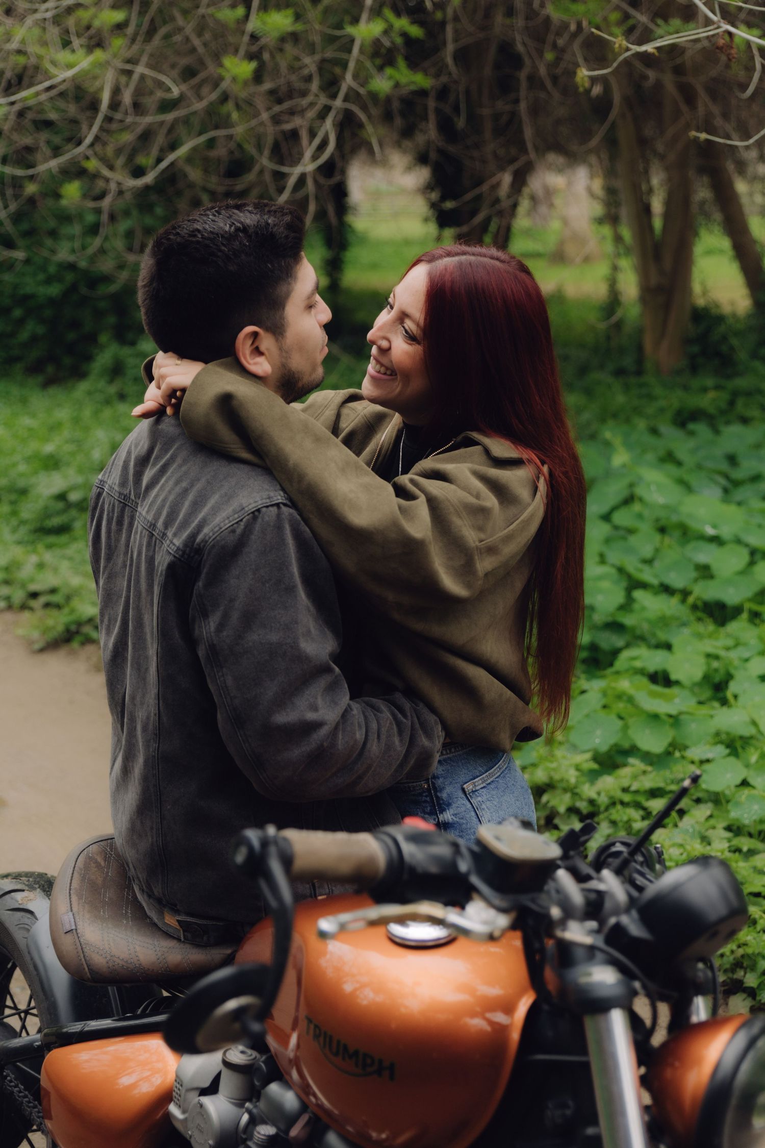 Couple sharing a tender moment together on a classic orange motorcycle in a green park.