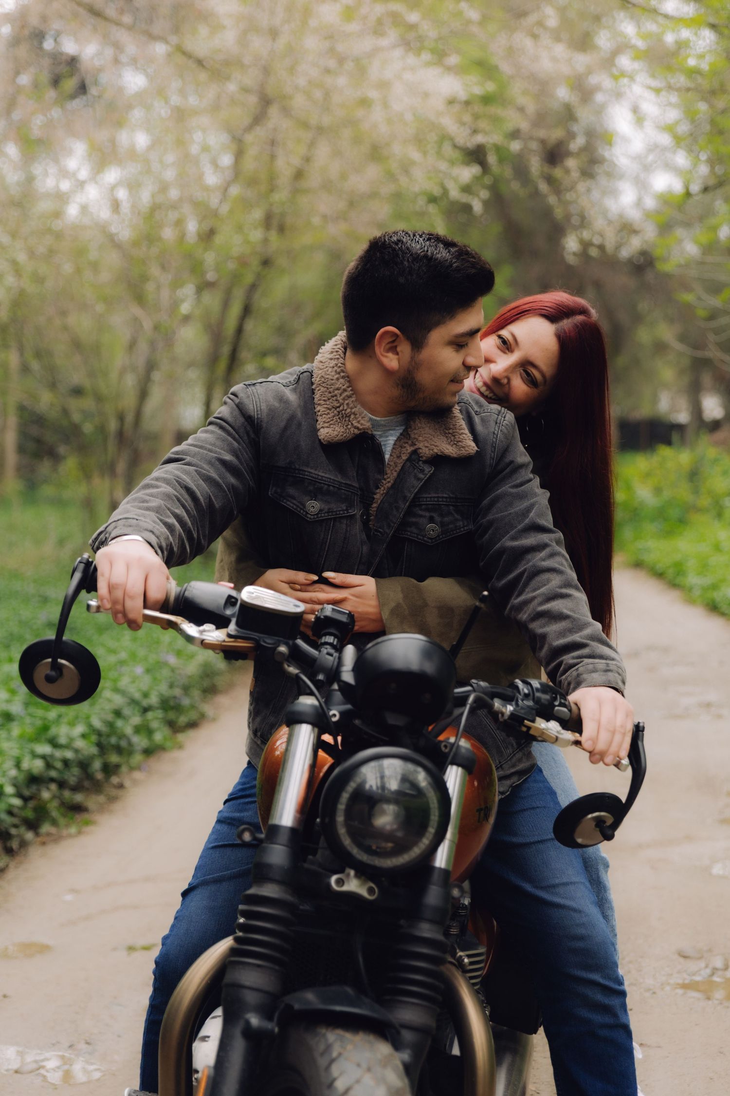 Smiling couple riding together on a vintage motorcycle along a scenic tree-lined path.