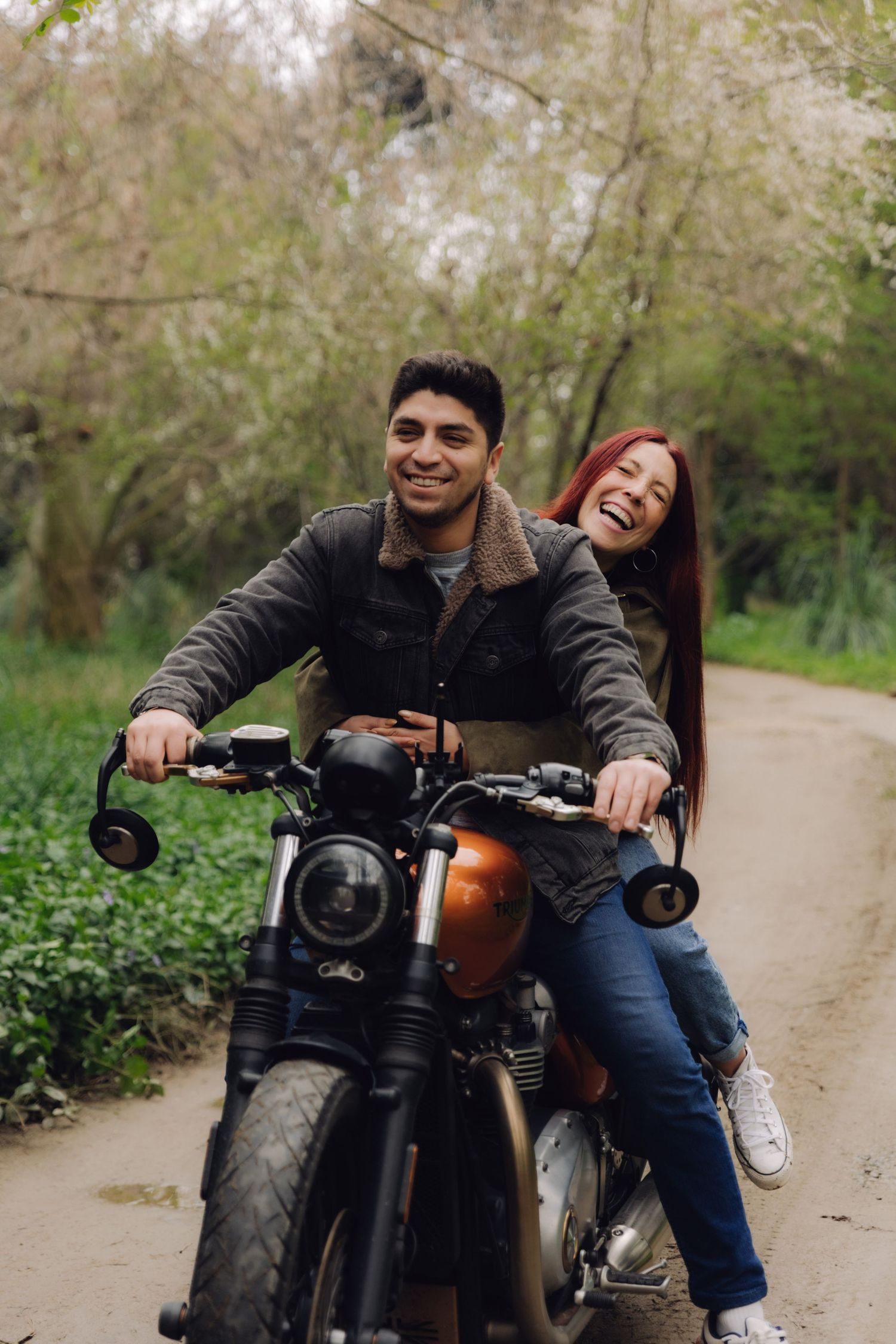 Happy couple laughing while riding a classic orange motorcycle through a lush green park.